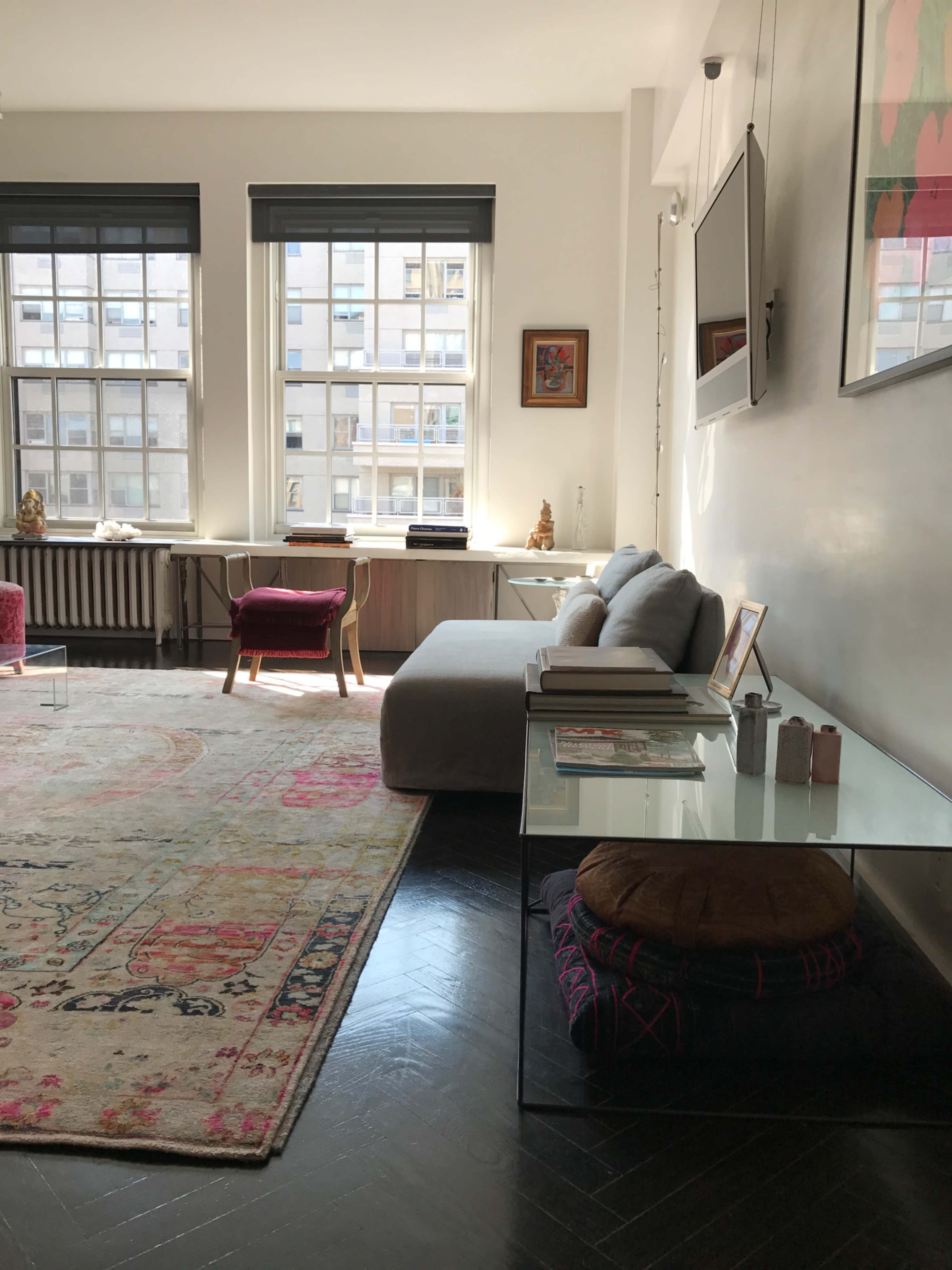A living room features a gray sofa, a glass side table, a colorful rug, and large windows with city views.