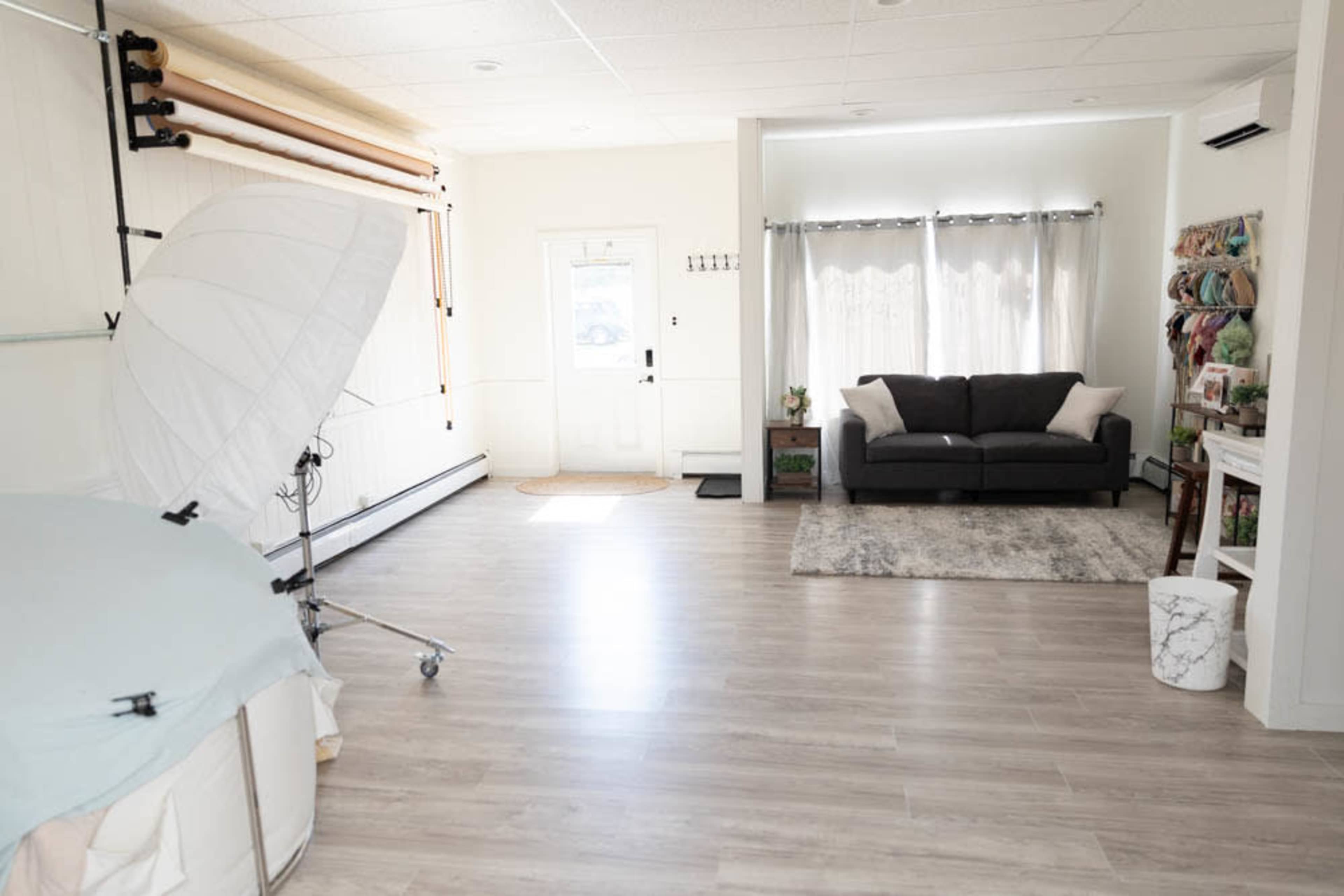 A spacious, well-lit room with a sofa, a large reflector umbrella, and a backdrop setup, featuring light-colored walls and wooden flooring.