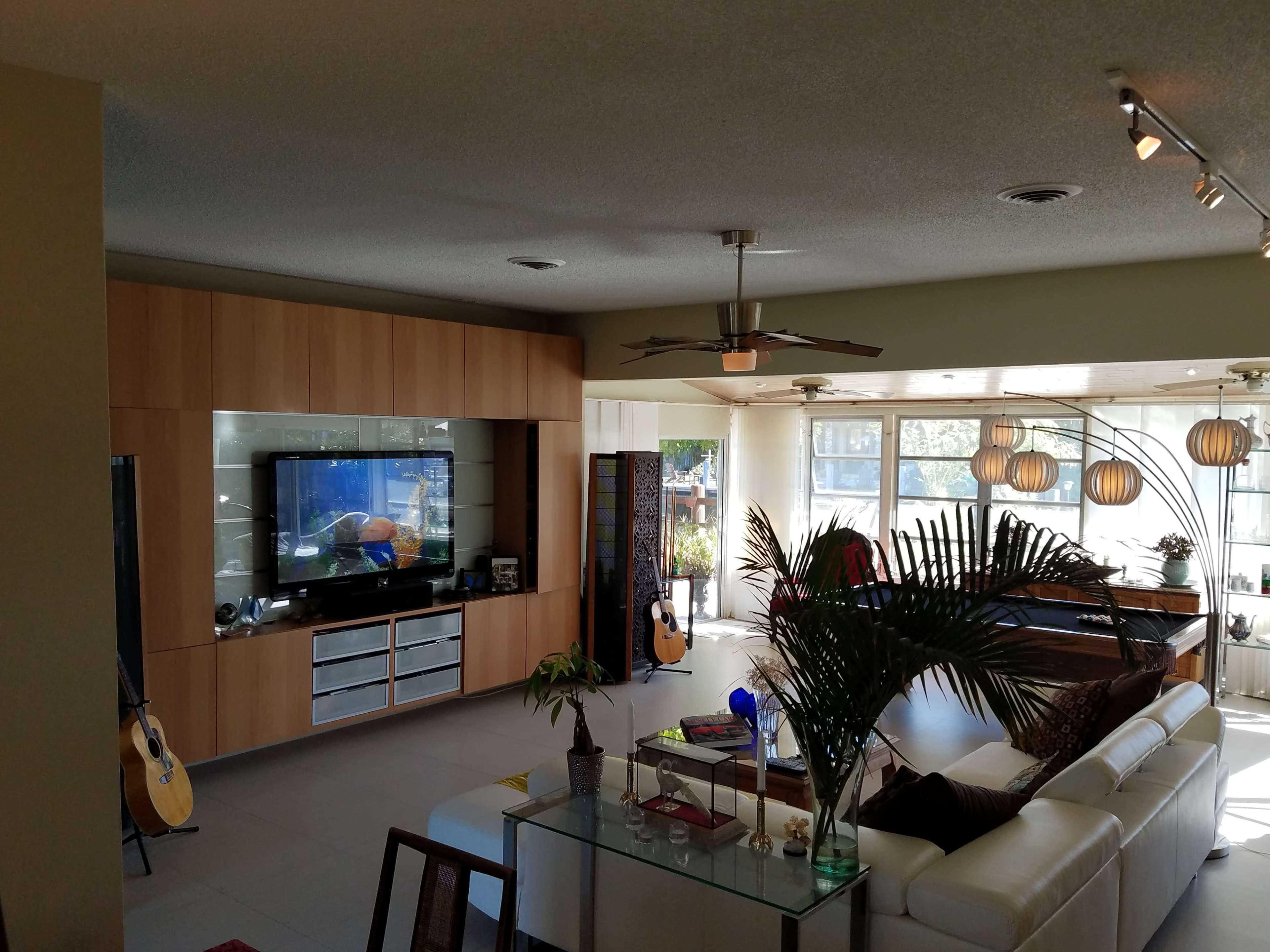 The scene shows a modern living room with a large sectional sofa, a wall-mounted television, and large windows allowing natural light to enter the space.