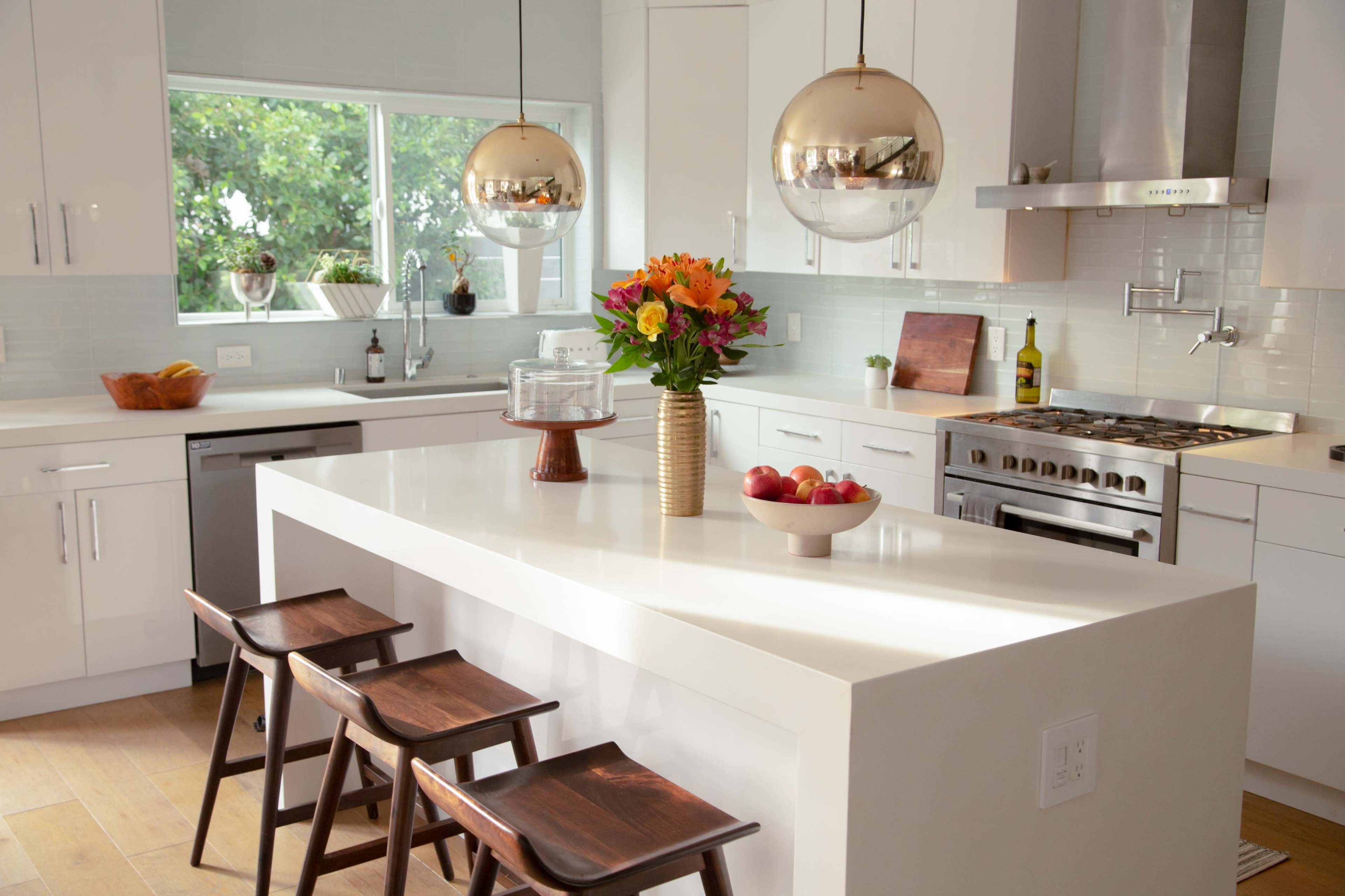 A modern kitchen features white cabinetry, a central island with three wooden stools, and pendant lighting above, adorned with a vase of flowers and a bowl of apples.