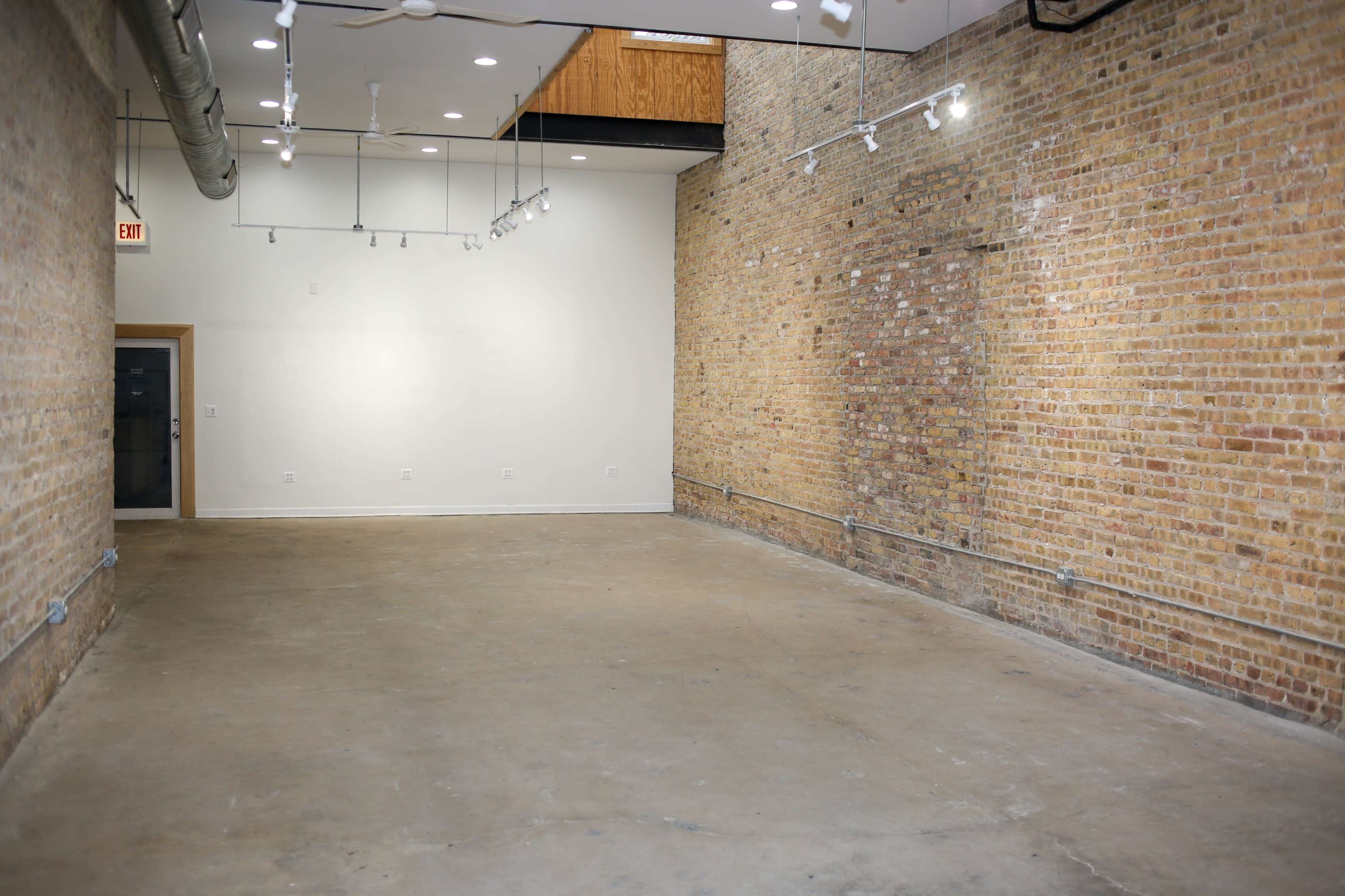 A spacious, empty room with exposed brick walls and concrete flooring, illuminated by track lighting.