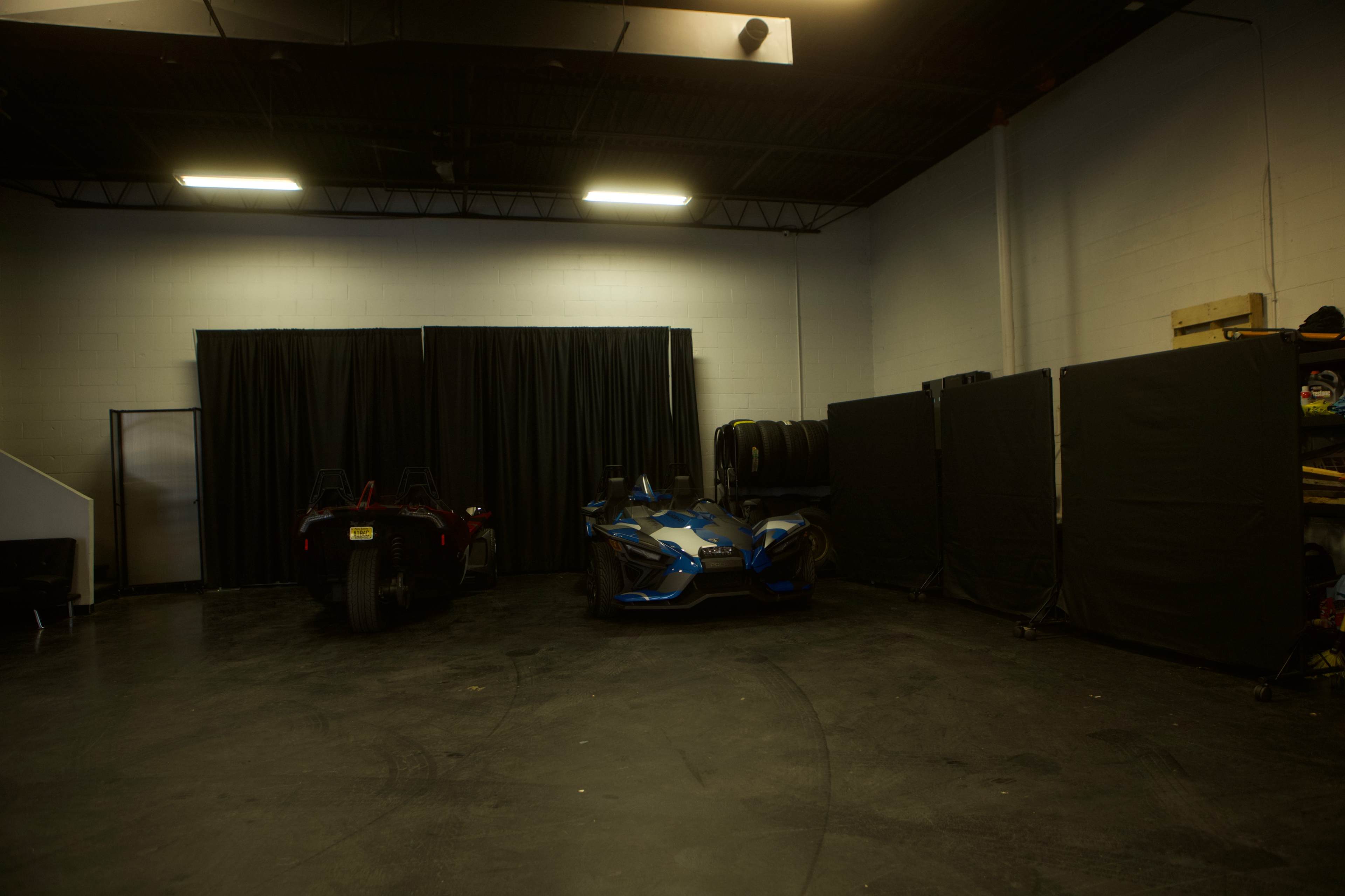 A dimly lit garage space with two vehicles, including a blue sports car, positioned near a black curtain and several tire stacks against the wall.
