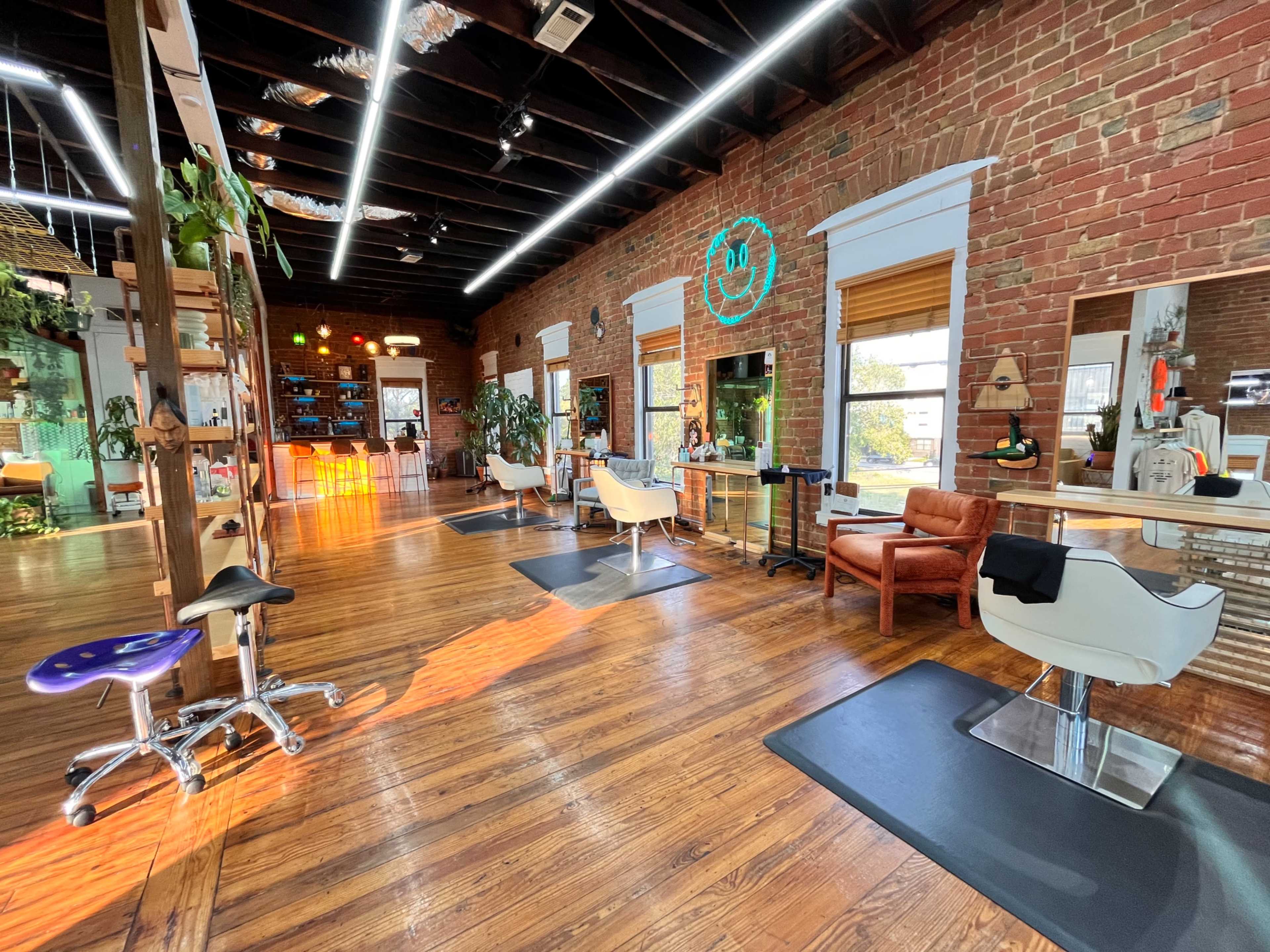 A hair salon with wooden floors, exposed brick walls, and styling stations equipped with mirrors and chairs, along with a waiting area featuring a leather chair.