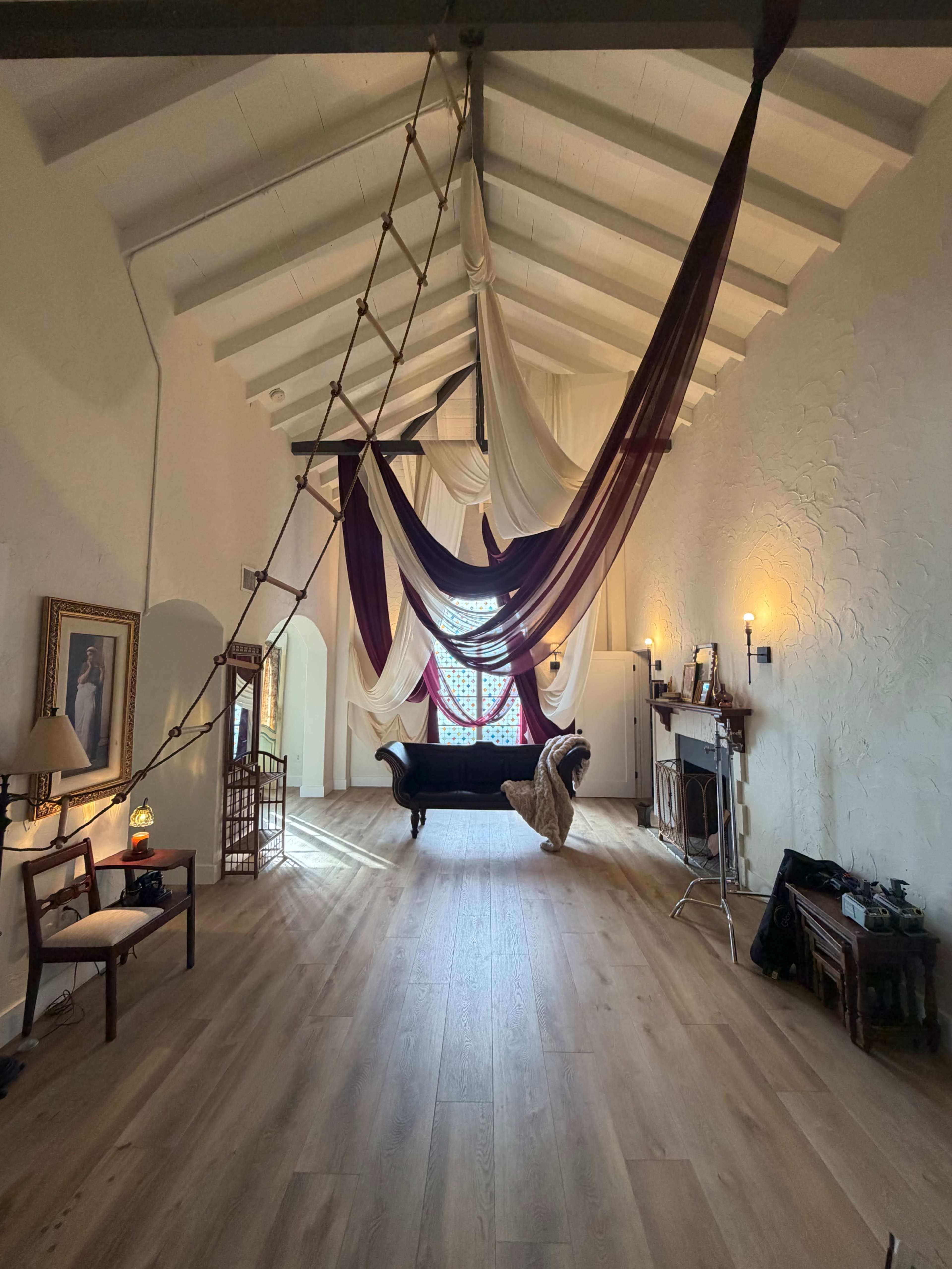 The image shows a spacious, well-lit room with a high ceiling, featuring draped fabric hanging from above and a black sofa in the center.