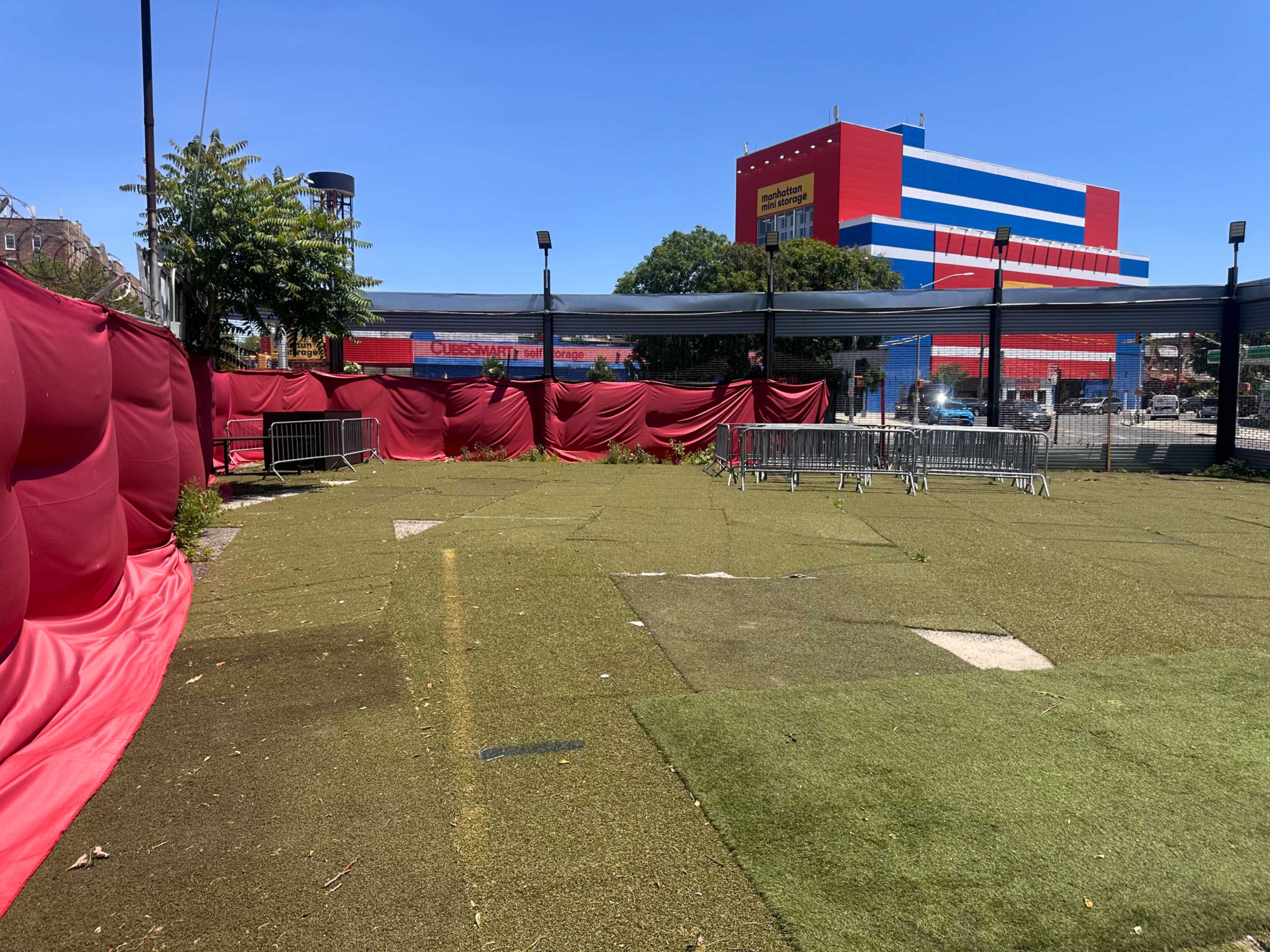 The image shows a vacant outdoor area with artificial grass, surrounded by red fabric drapes and metal barriers, under a clear blue sky.