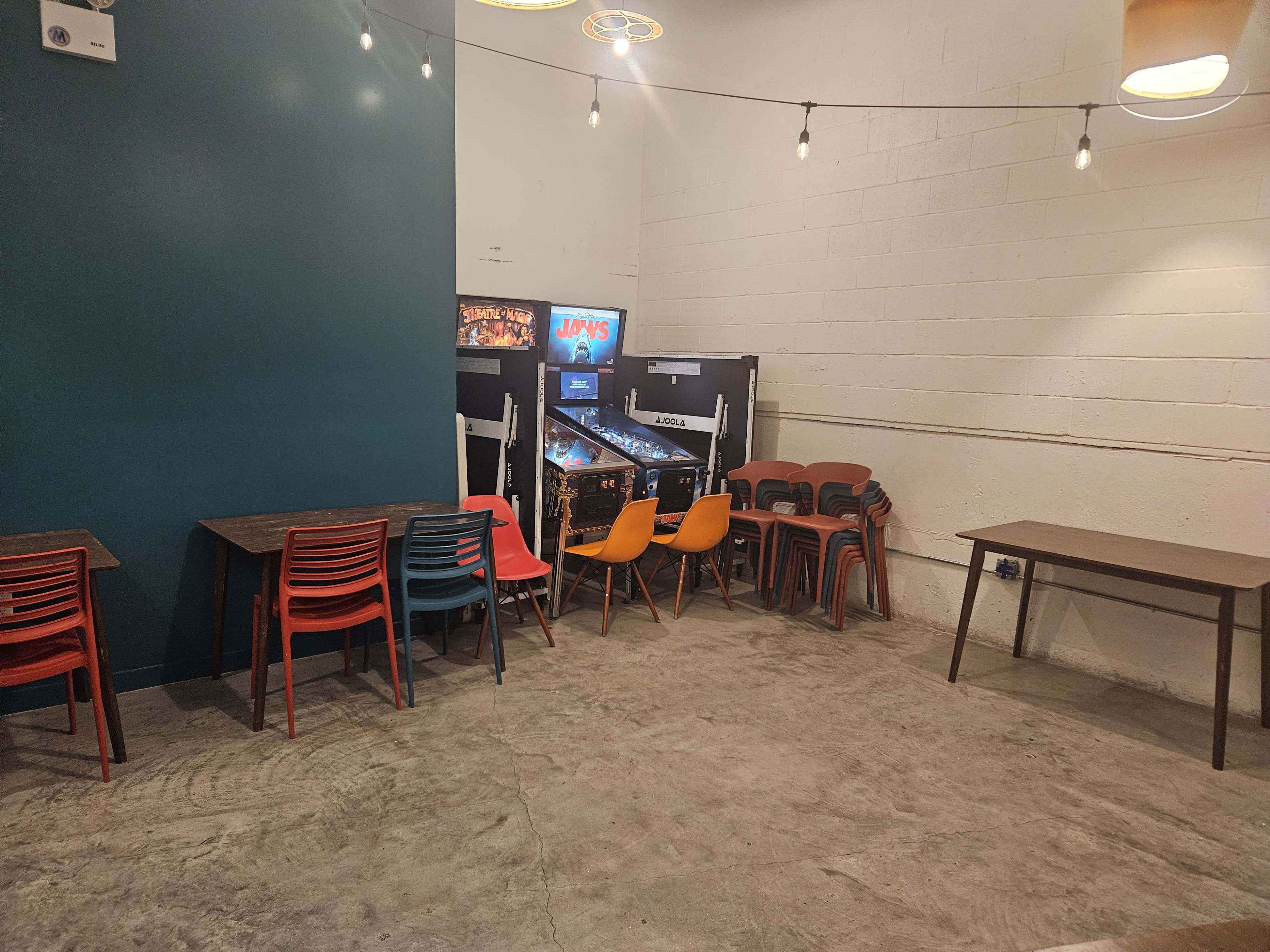 The image shows a seating area with wooden tables and colorful plastic chairs, alongside two arcade machines in a dimly lit space.