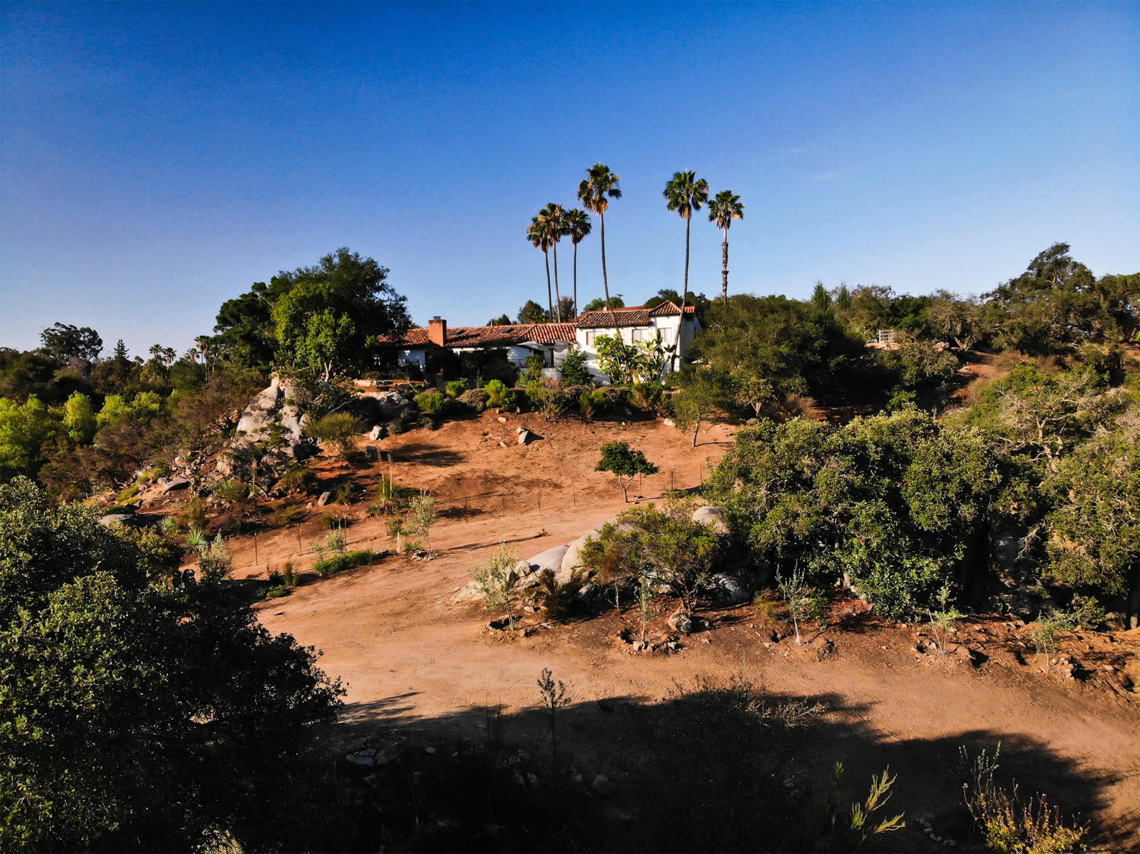 A white house sits on a hillside surrounded by various trees and shrubs under a clear blue sky.