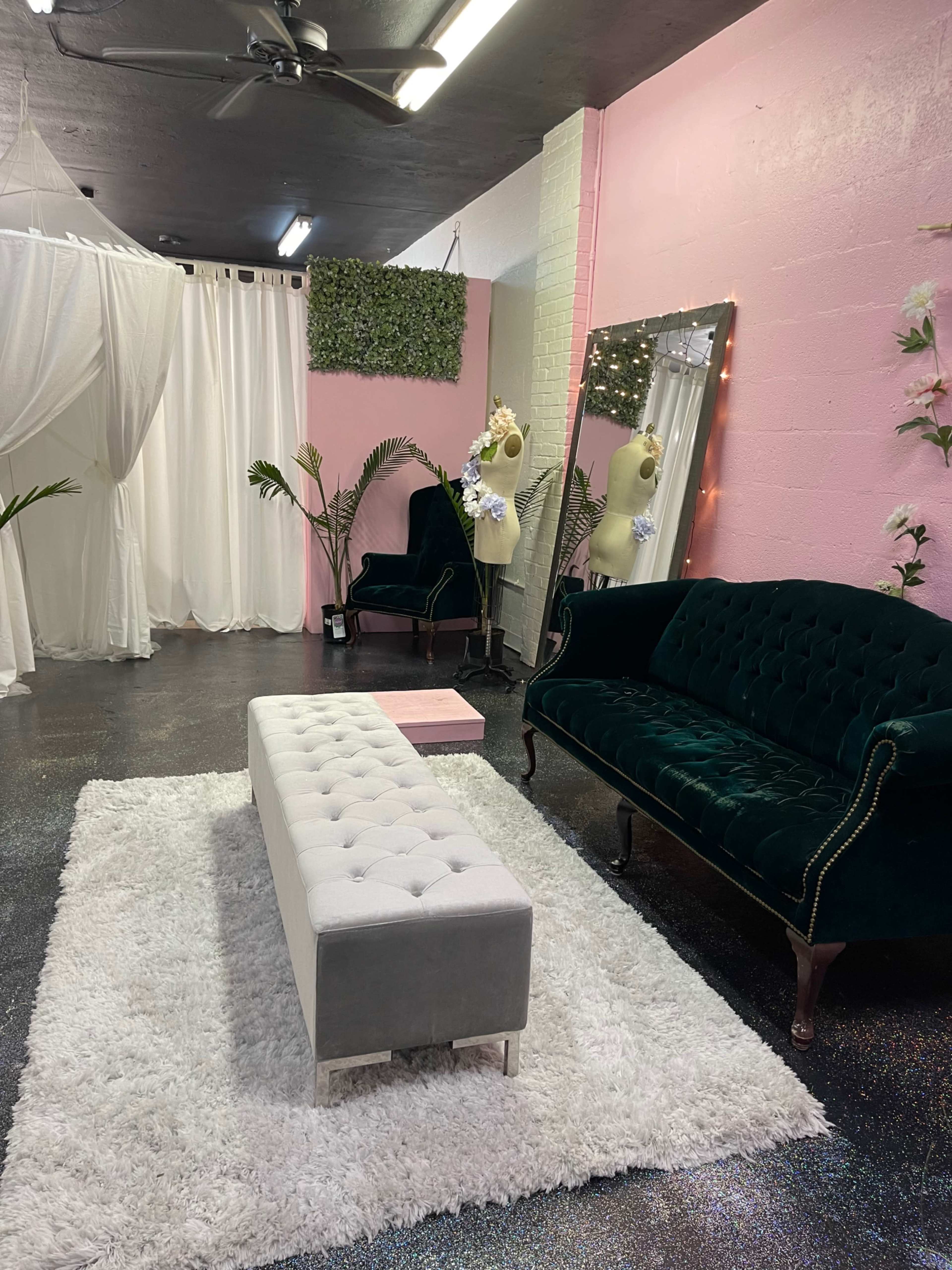 A pink and white interior space featuring a green velvet sofa, a tufted gray ottoman, and plants, with a decorative mirror and white drapes.