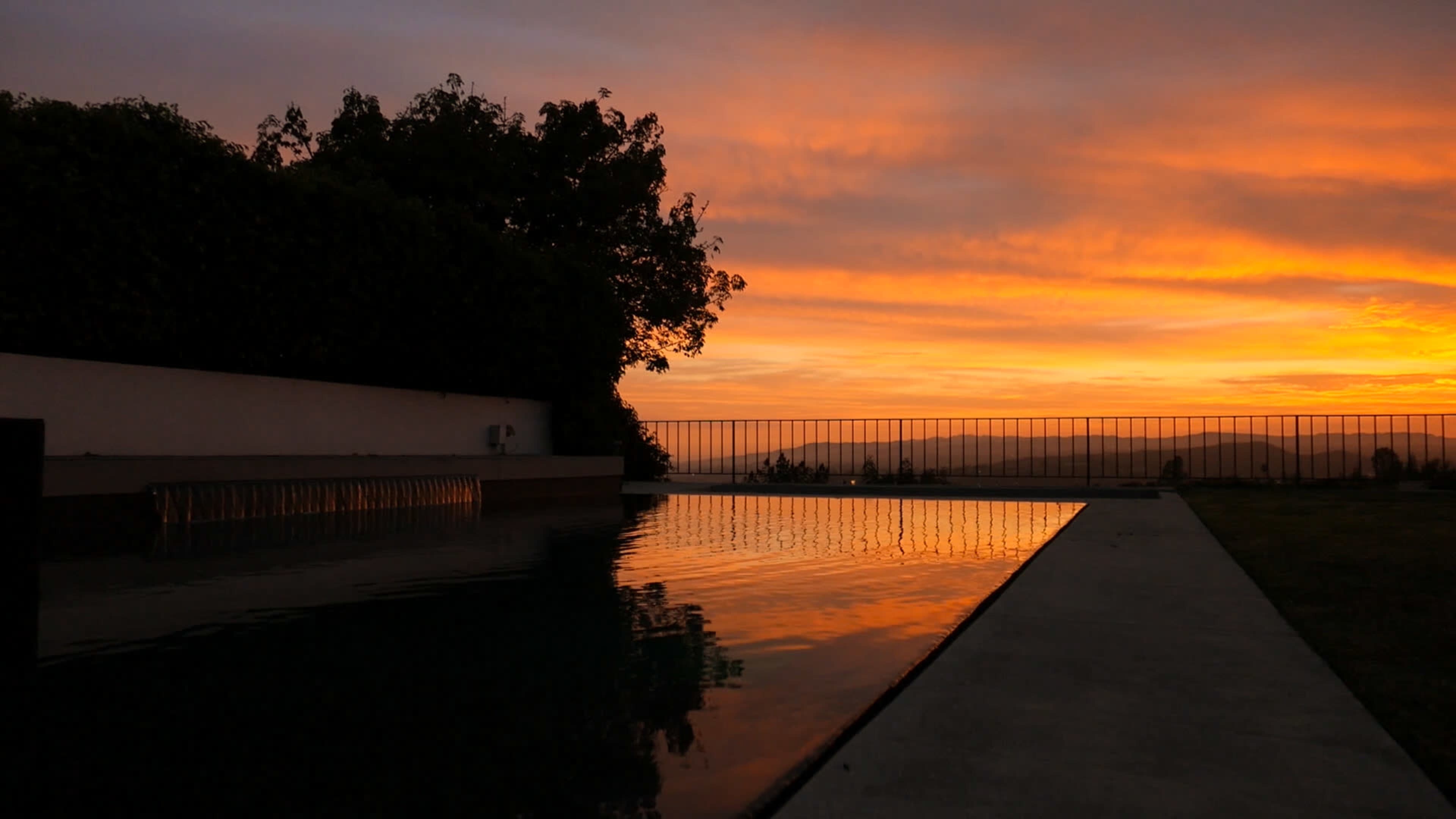 A calm swimming pool reflects a vibrant sunset sky, framed by greenery and a boundary fence.