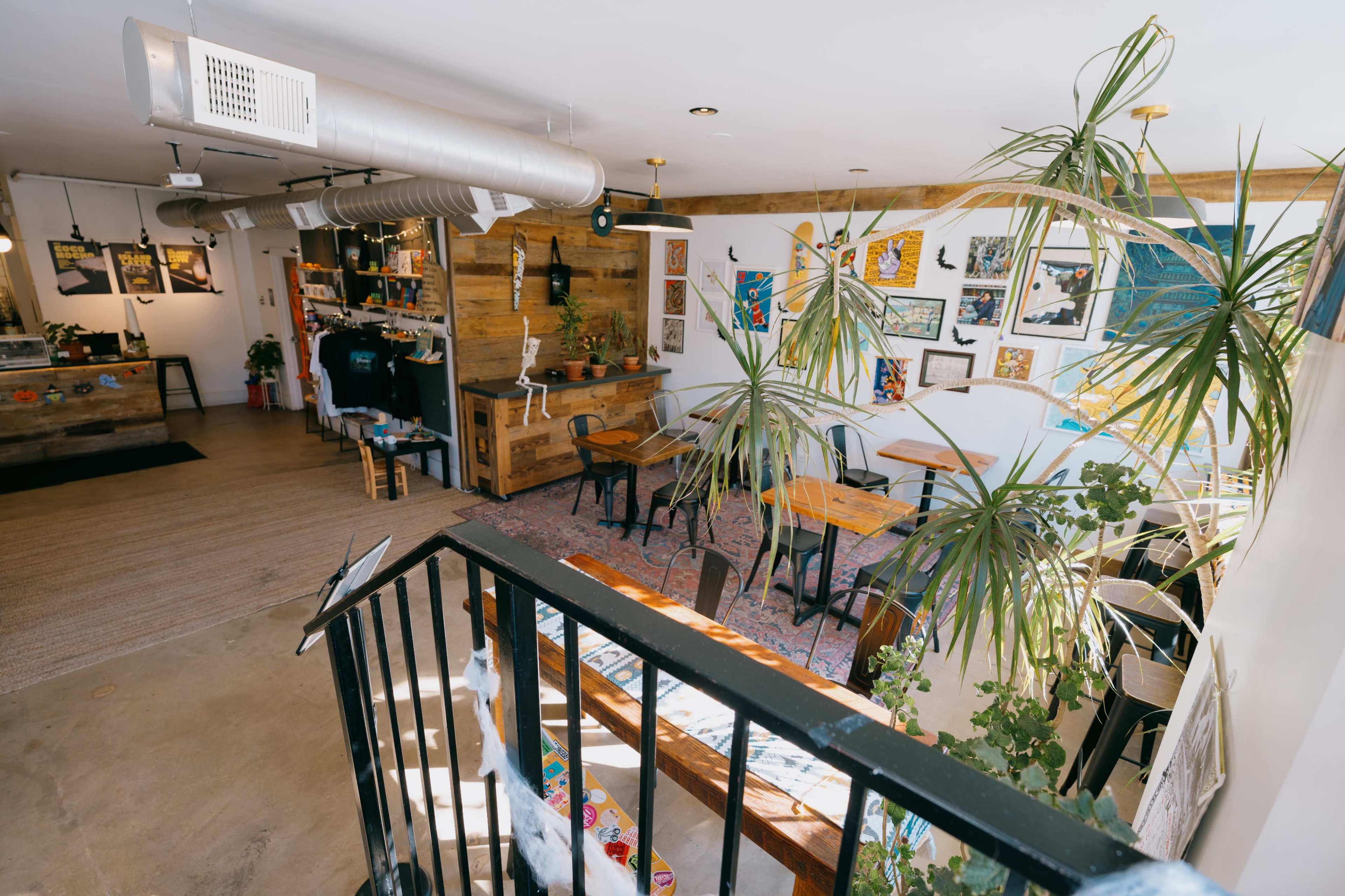 The image shows a cozy café interior with wooden tables, colorful artwork on the walls, and greenery throughout the space.