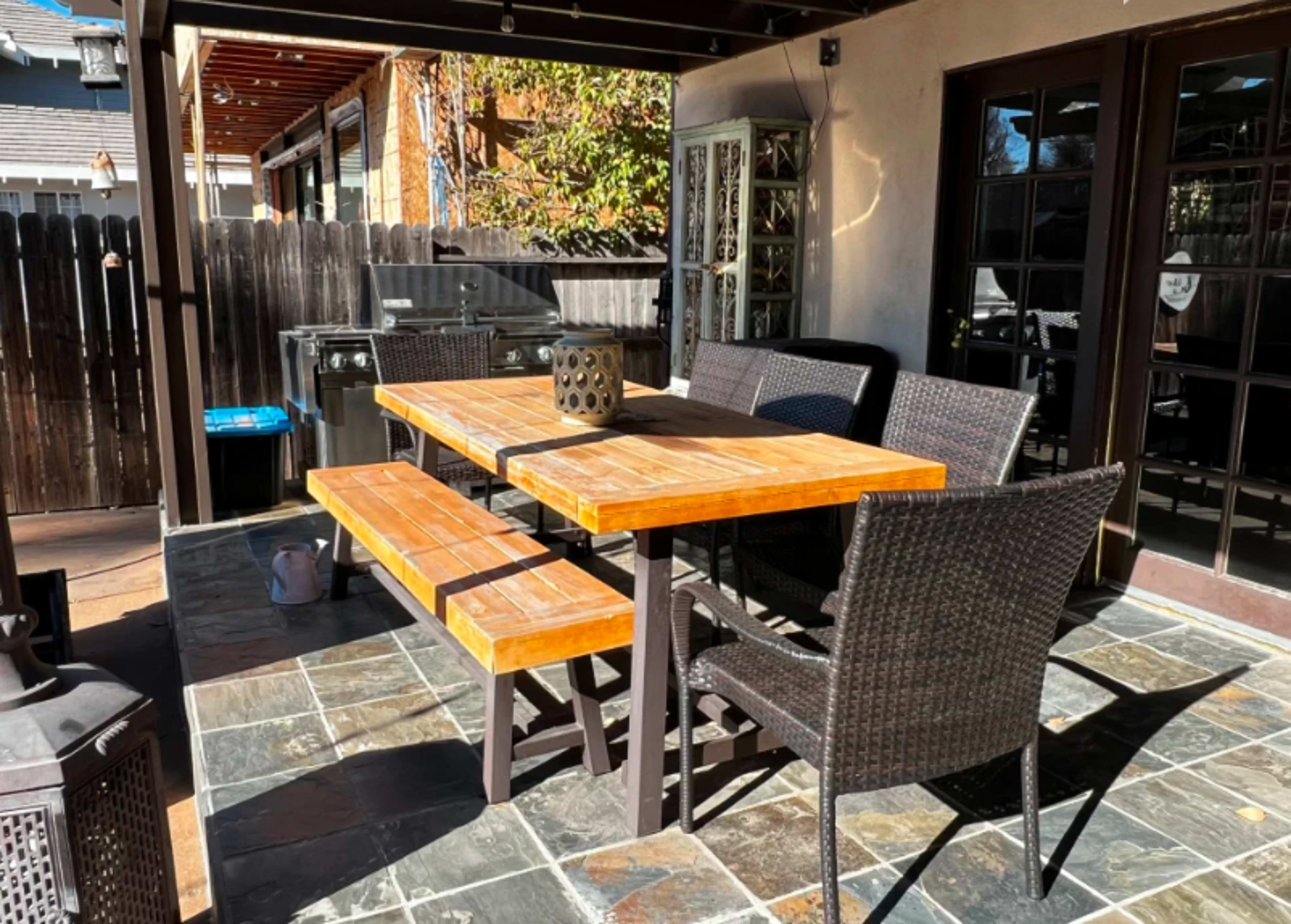A wooden dining table with benches and chairs is set under a covered patio next to a grill and sliding glass doors.