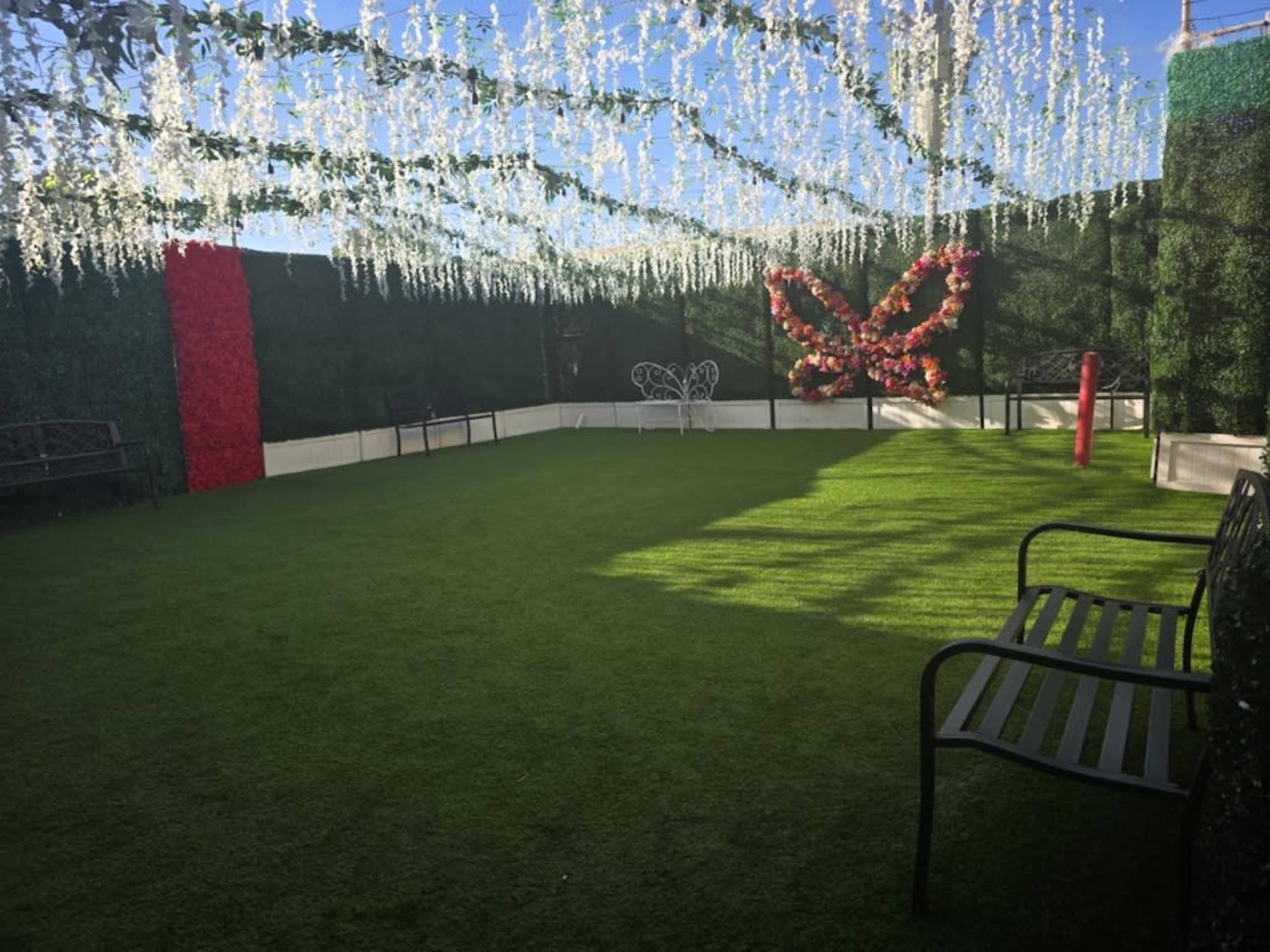 The scene features a landscaped outdoor area with artificial grass, adorned with floral decorations and a large butterfly installation against a backdrop of greenery.