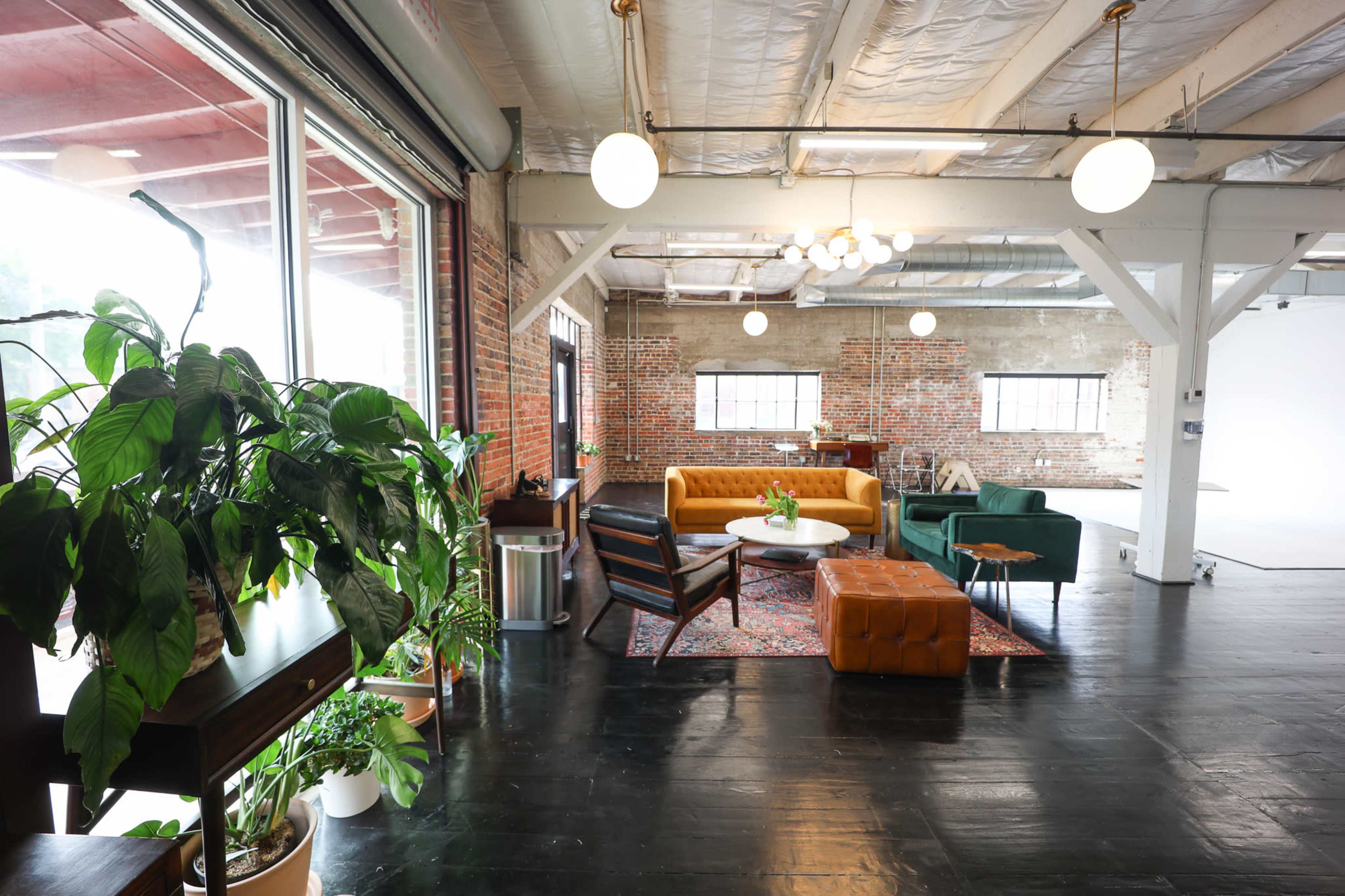 The image shows a spacious, modern interior with comfortable seating arrangements, large windows, and various green plants.