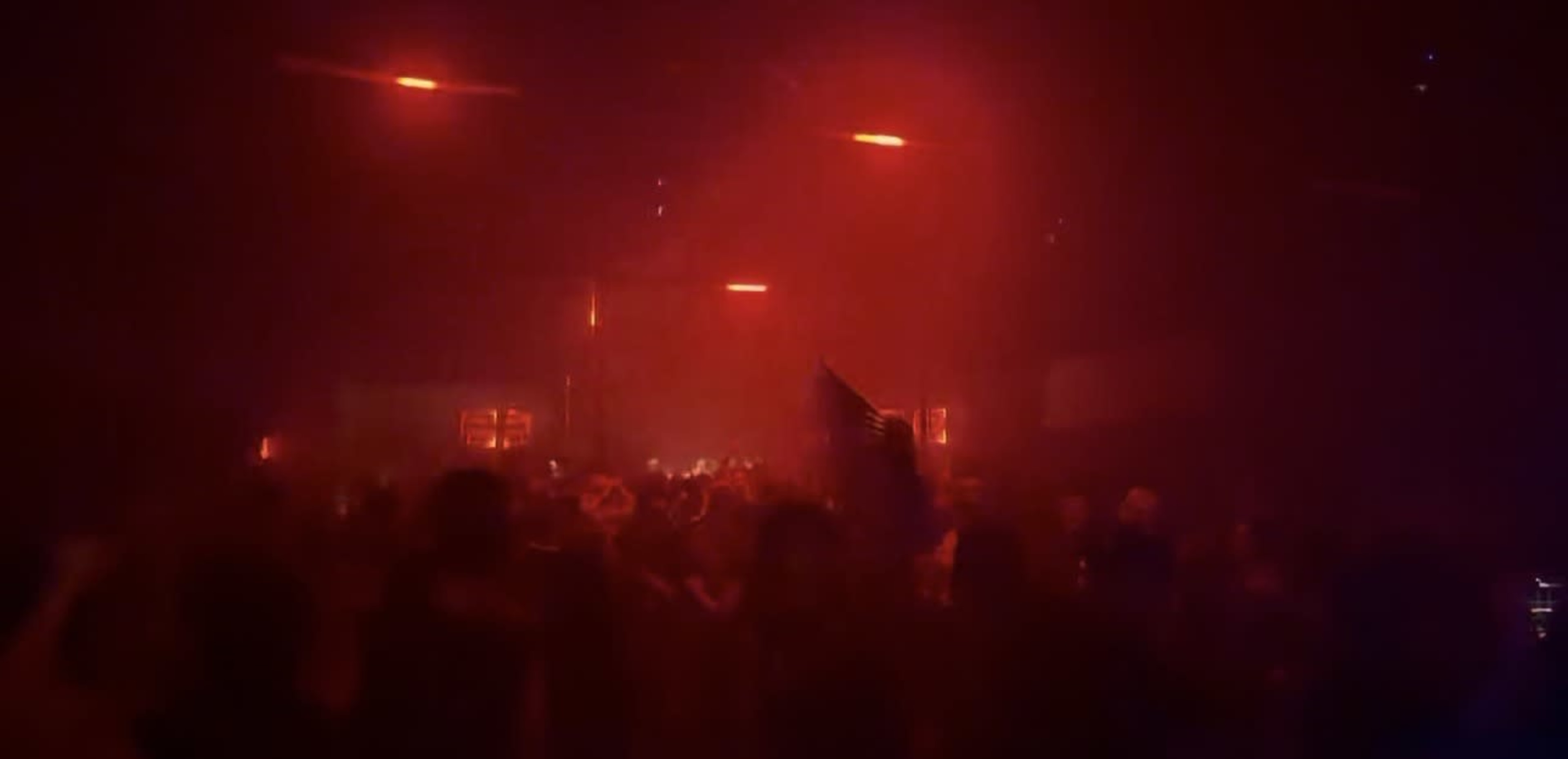 A crowd gathers in a dimly lit venue with red lighting and smoke filling the air.