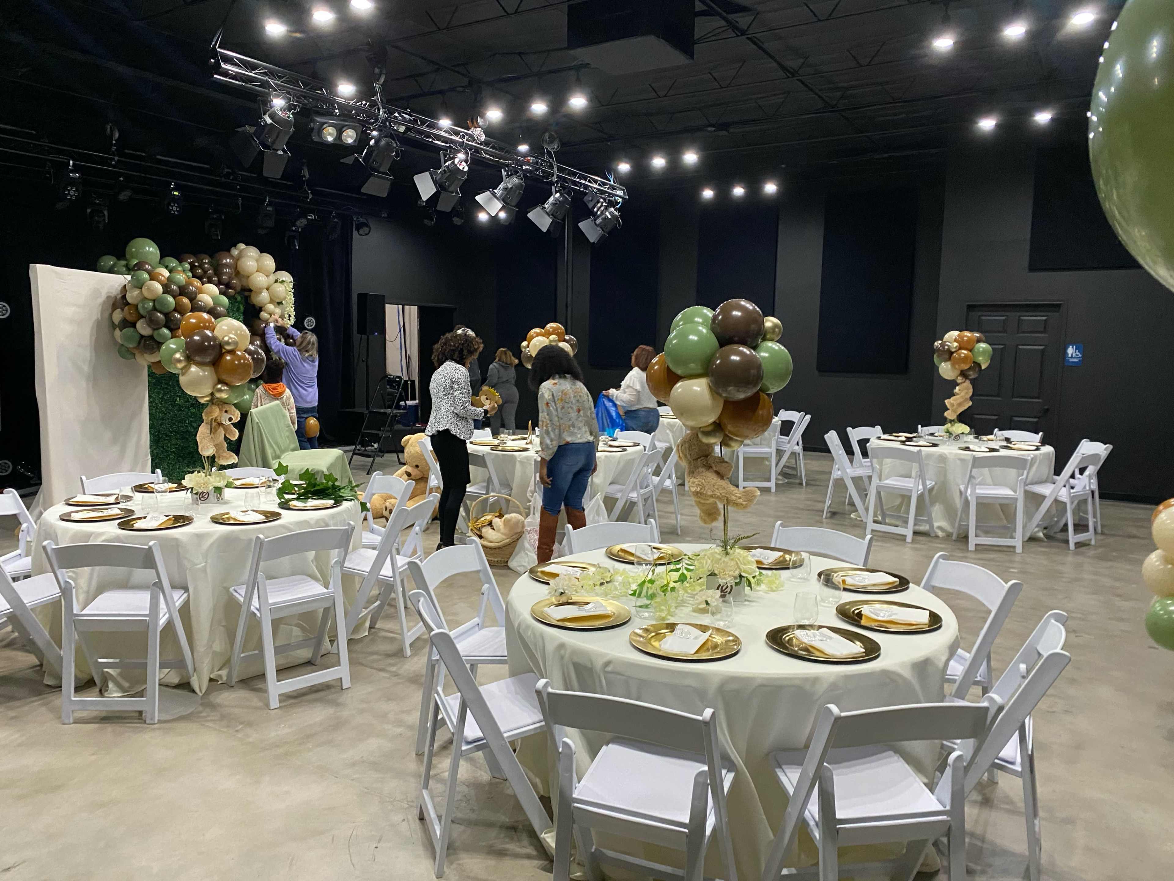 The image shows a decorated event space with round tables set with plates and centerpieces, surrounded by balloon arrangements and attendees preparing for the event.