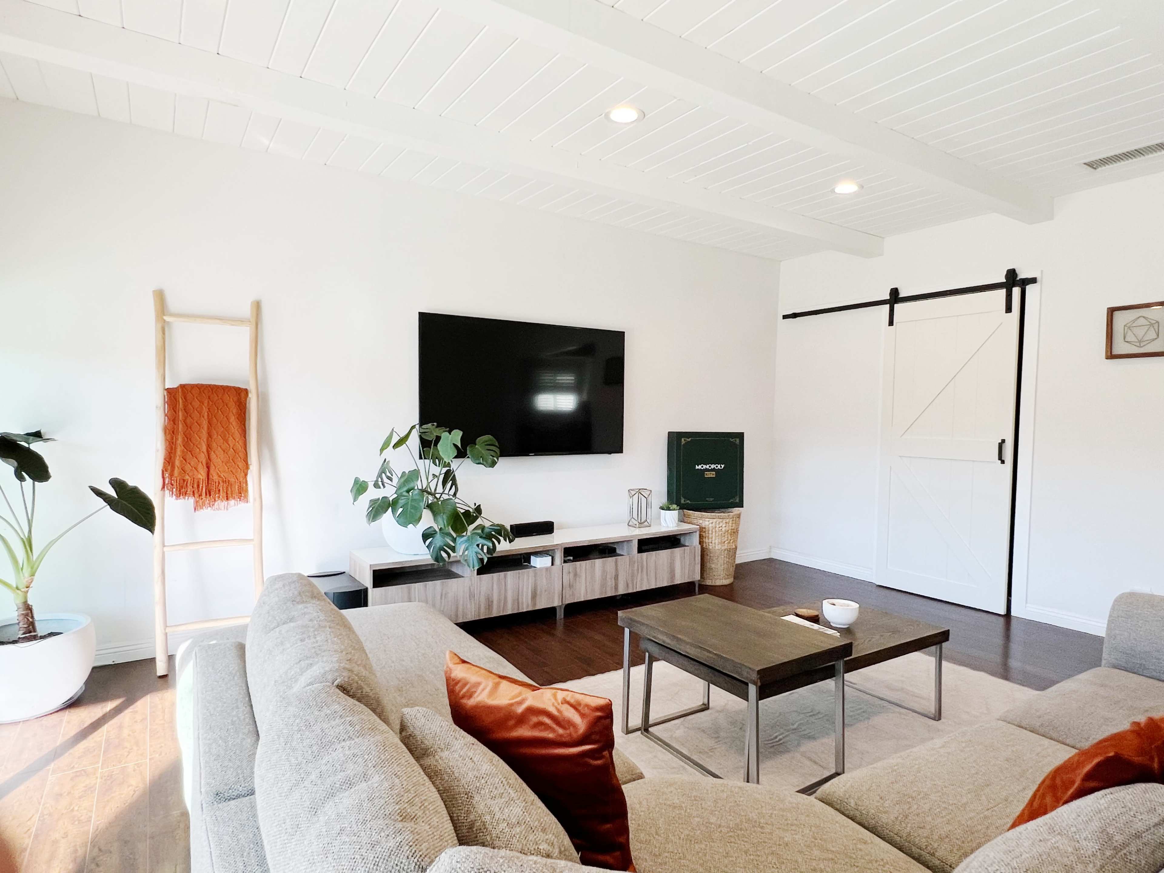 The living room features a sectional sofa, a coffee table, a mounted television, and a sliding barn door, with decorative plants and a cozy throw.