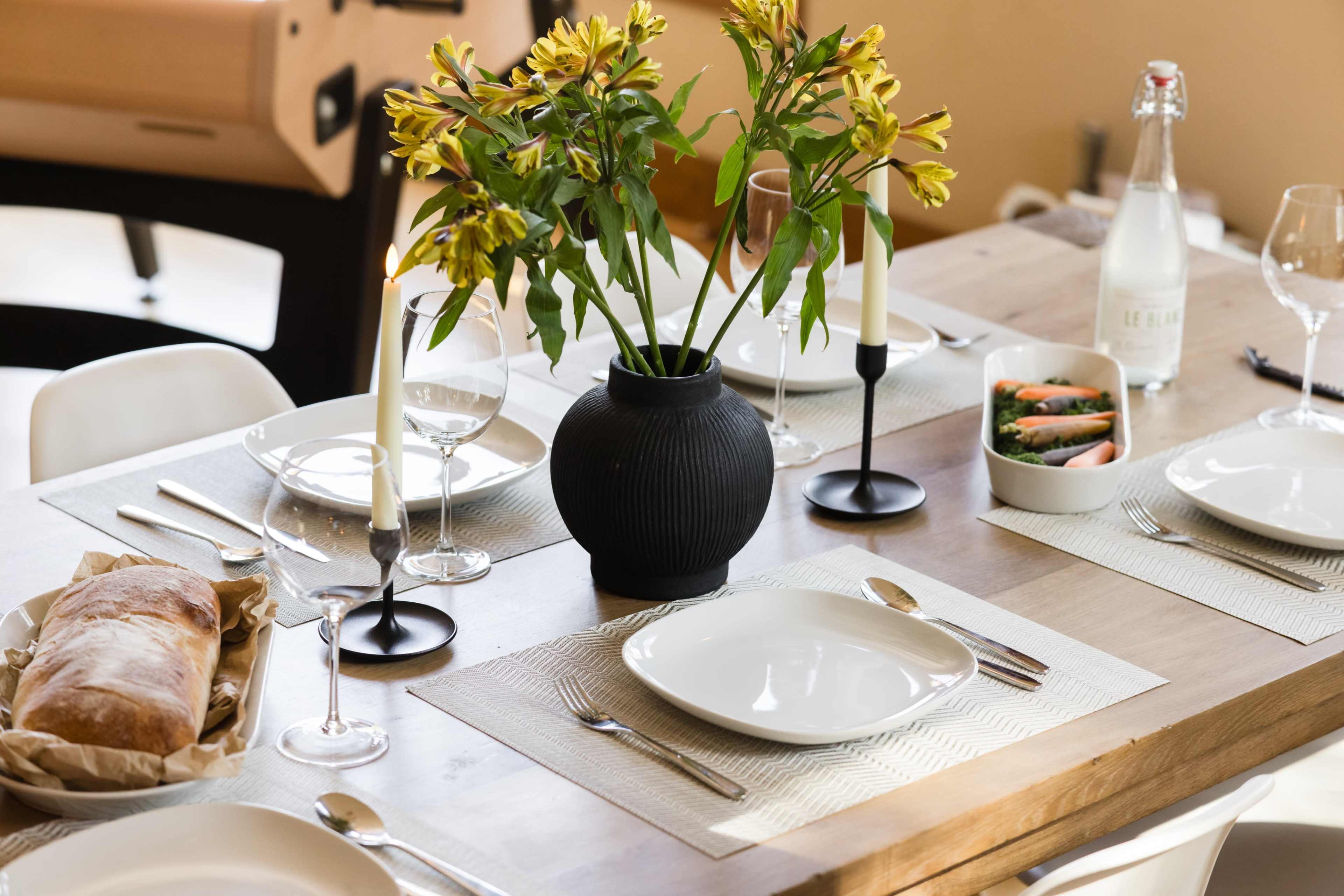 A neatly arranged dining table features white plates, cutlery, glasses, and a vase of yellow flowers, with bread and a dish of vegetables on the side.