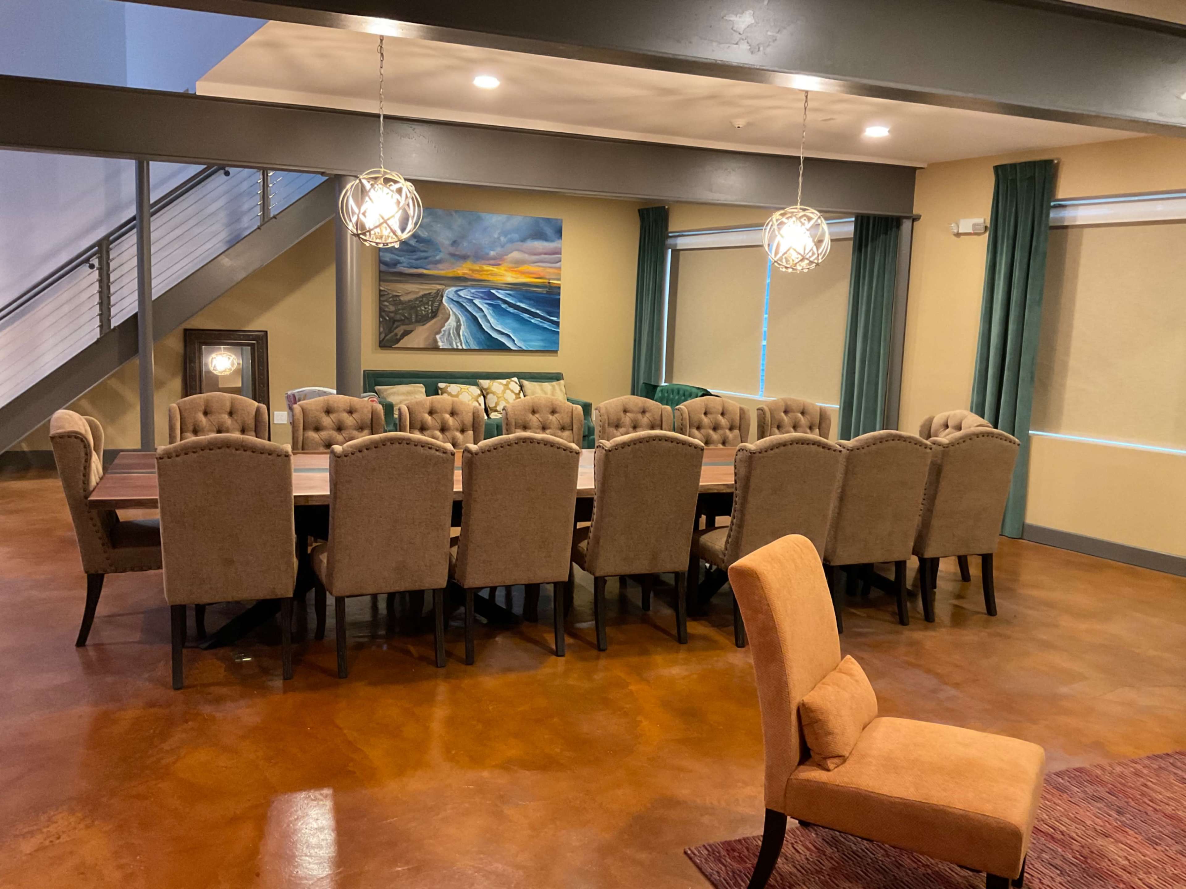 A large dining area with a long wooden table surrounded by upholstered chairs, a landscape painting on the wall, and stylish light fixtures overhead.