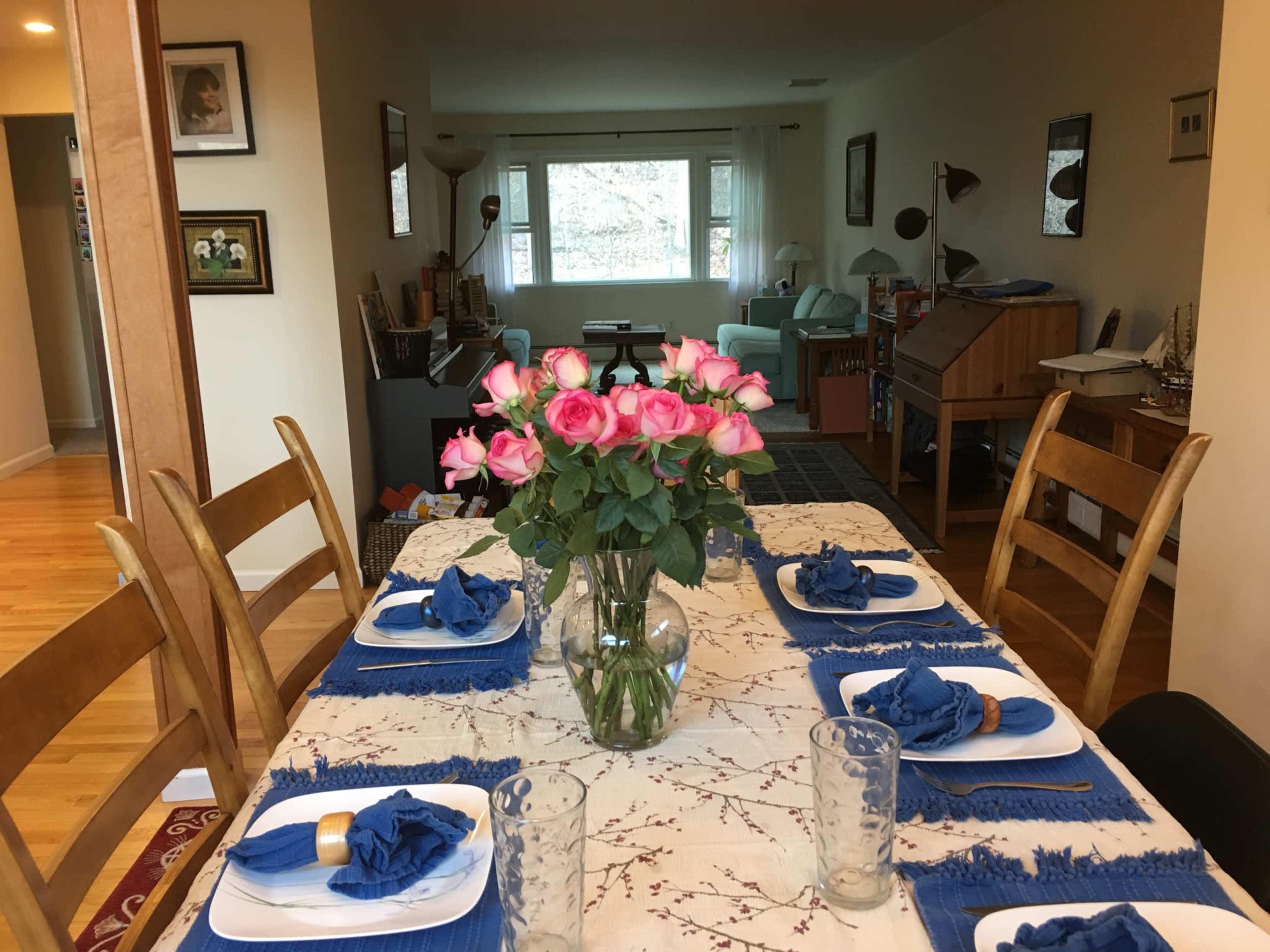 A dining table set with blue napkins and plates is adorned with a vase of pink roses, while a living room with light-colored furniture is visible in the background.