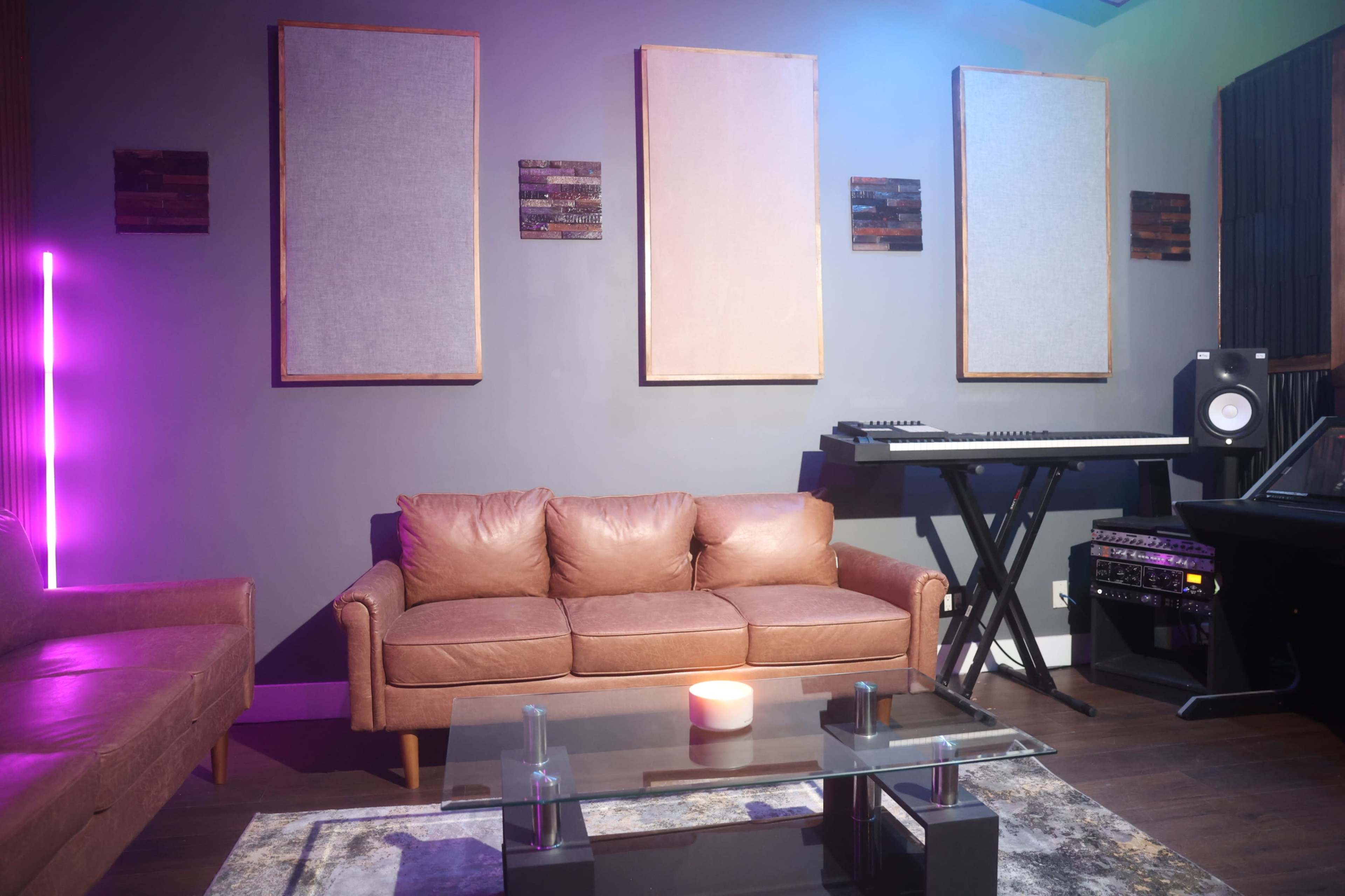 A contemporary lounge area features a leather sofa, a glass coffee table, and a music keyboard setup against a wall with three framed acoustic panels.
