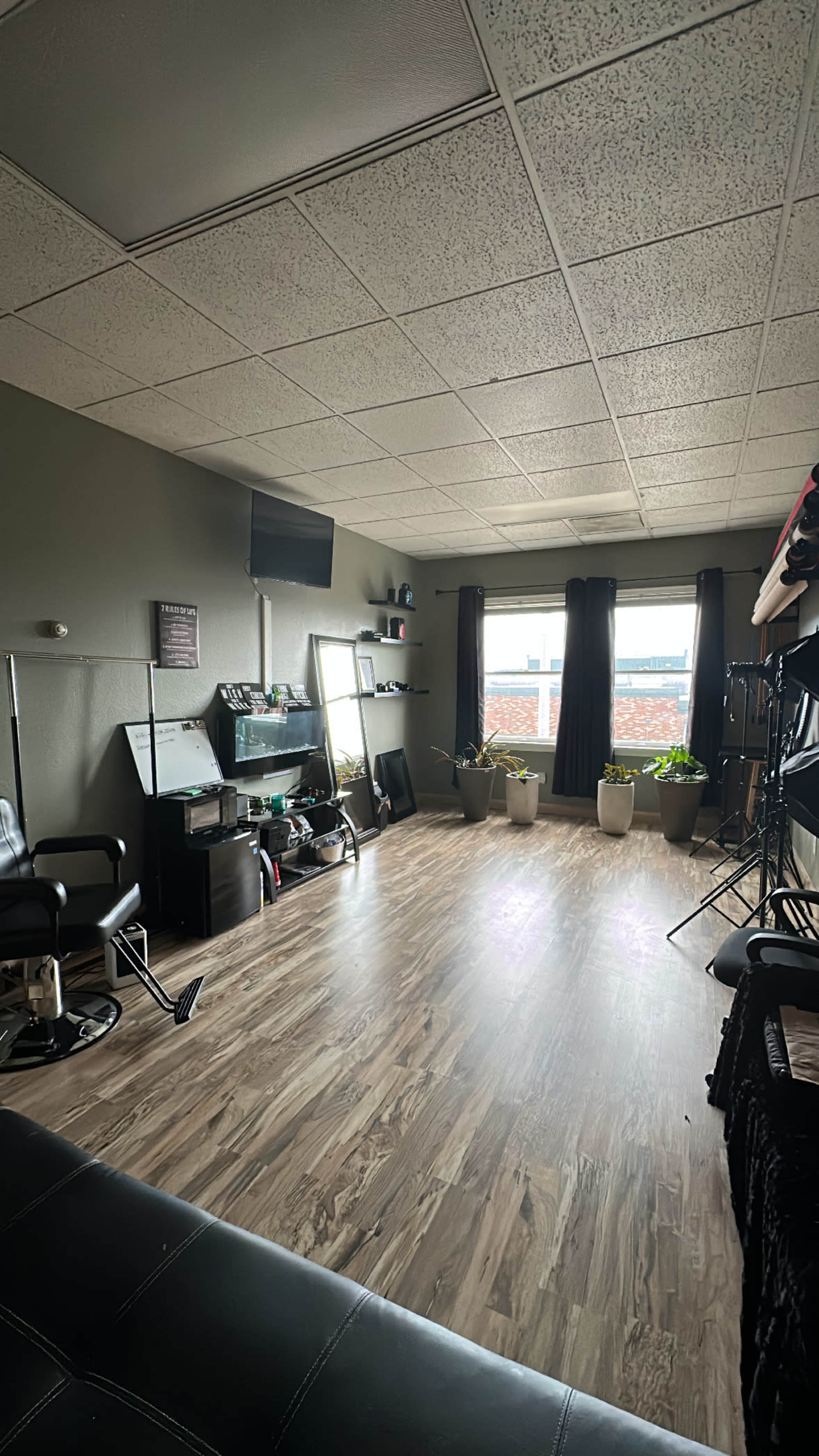 The image shows a spacious room with a wooden floor, large windows, several potted plants, and salon chairs along the walls.