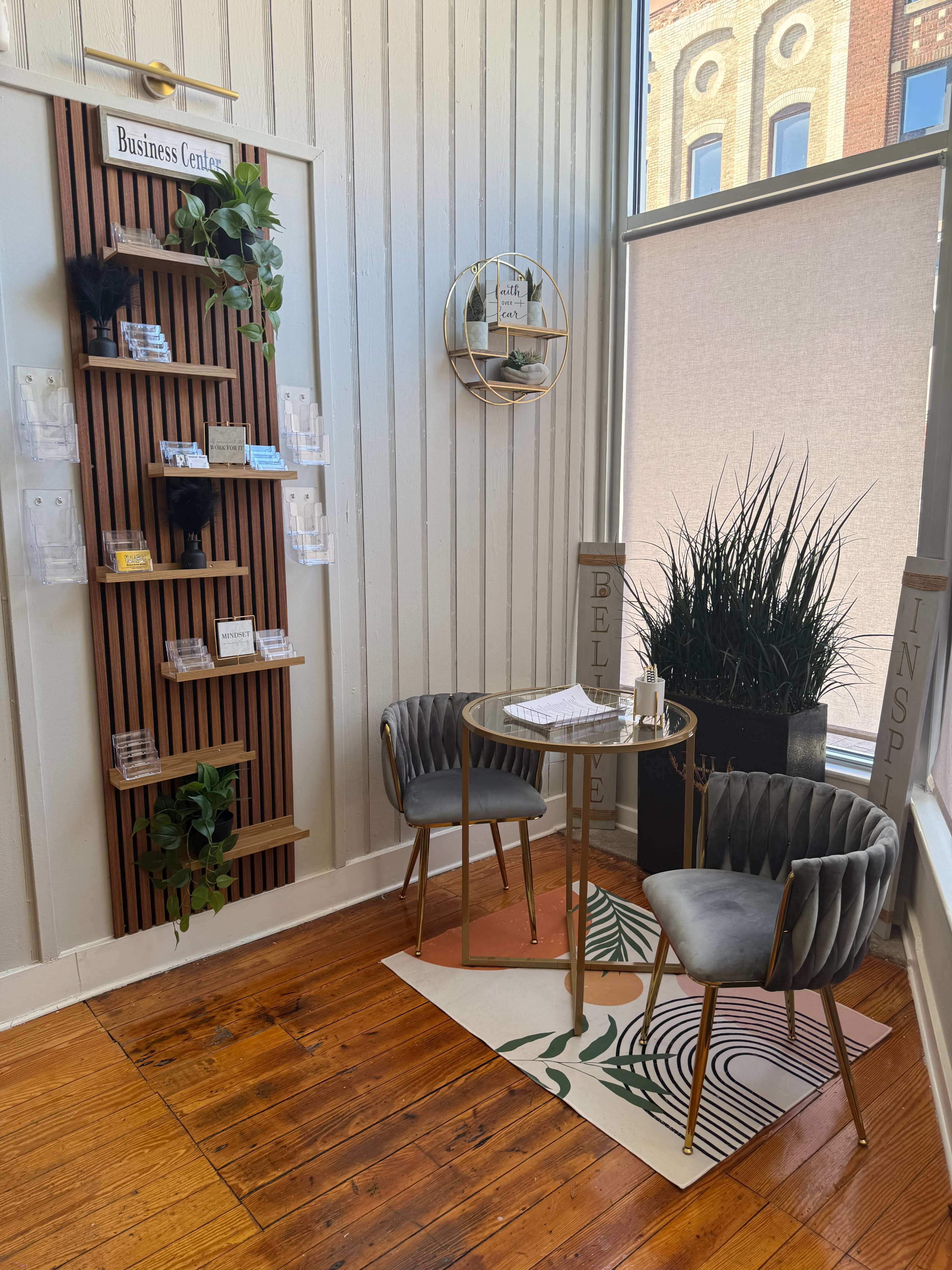 The image shows a small business center area featuring a wooden display for business cards, two upholstered chairs, a round table, and a potted plant in a corner.