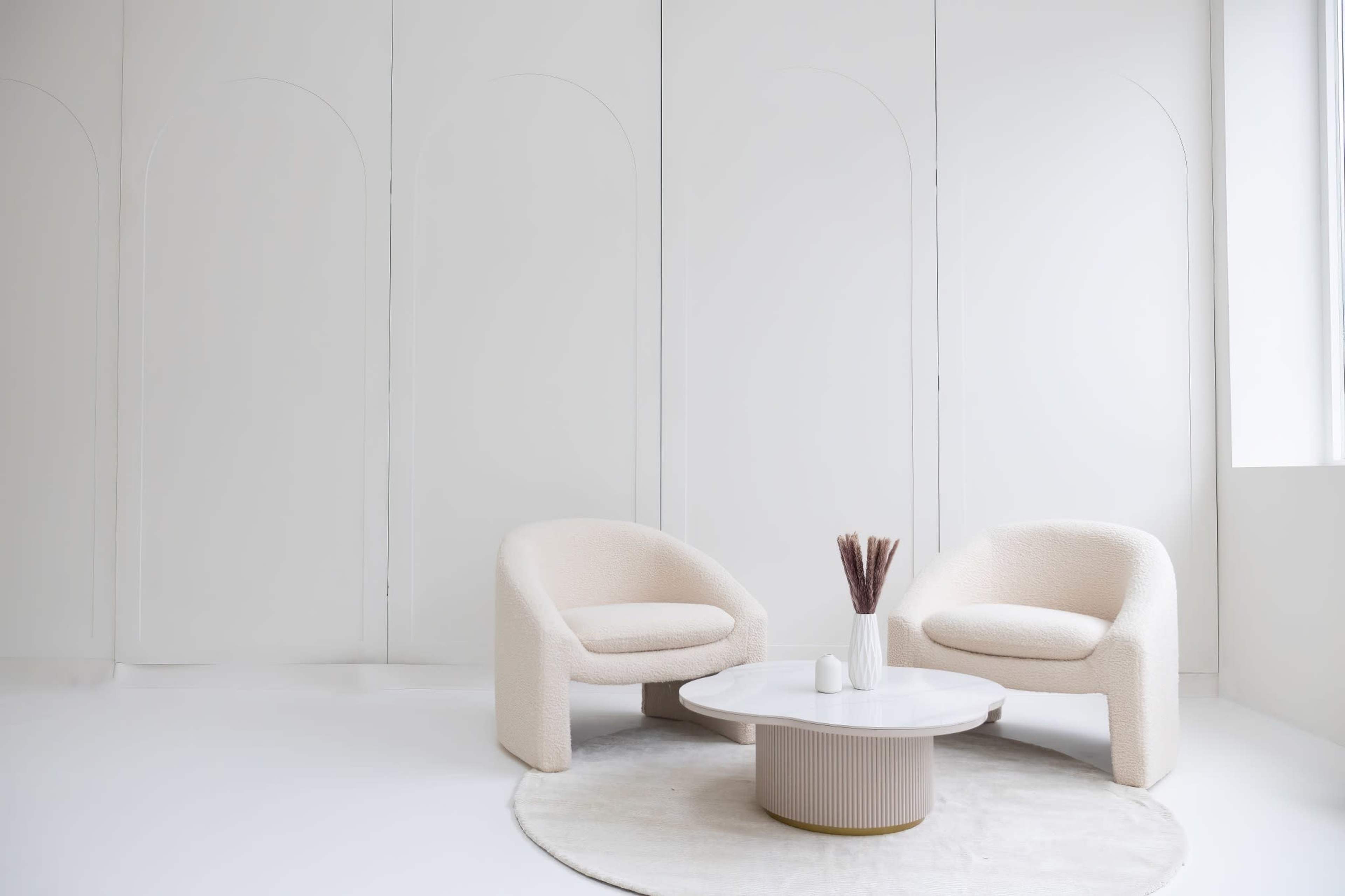 Two textured beige chairs facing a round white coffee table on a circular rug in a minimalist, bright room with white walls.