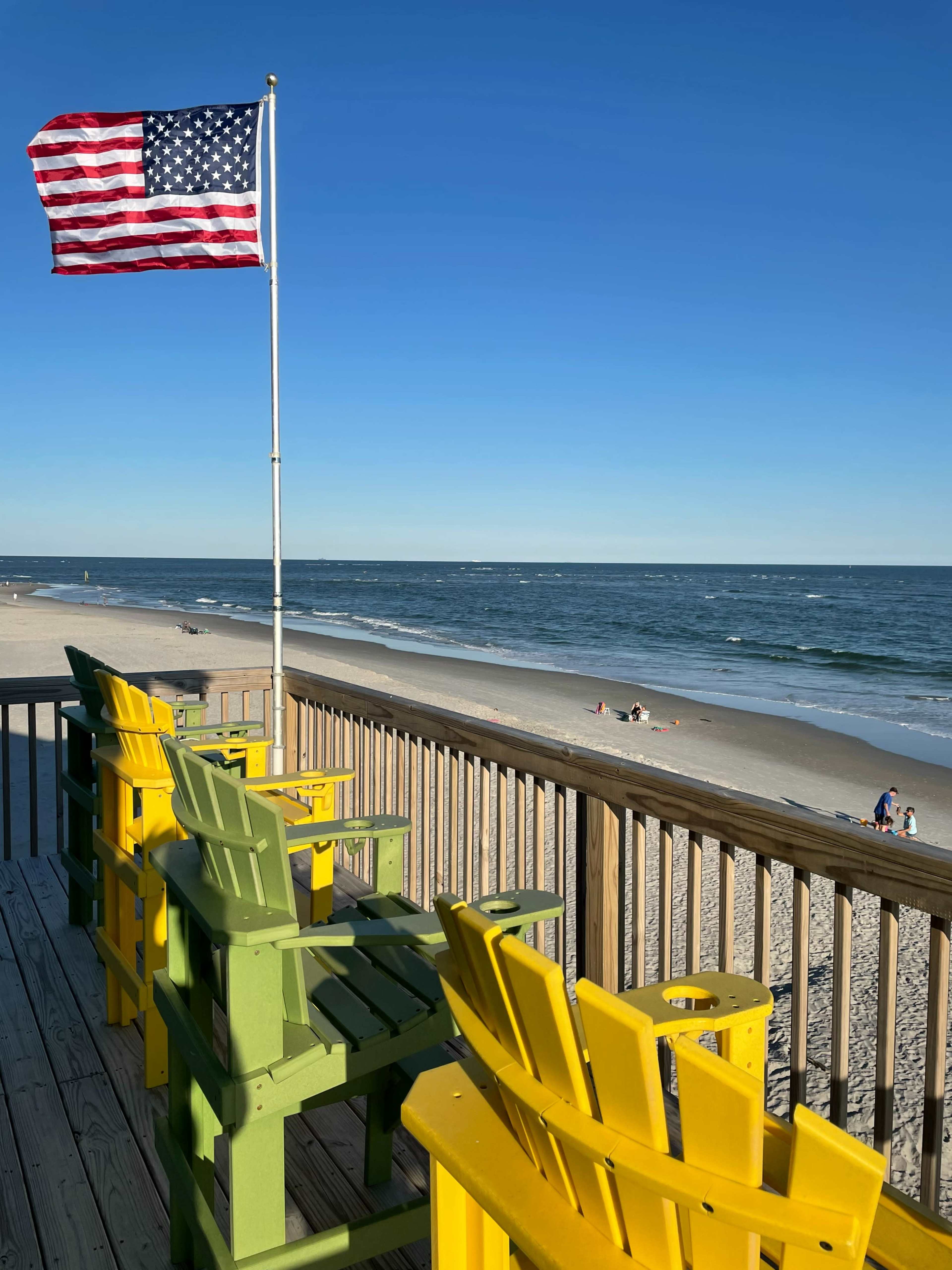 A flag of the United States flies next to colorful chairs on a deck overlooking a beach with gentle waves and a few people in the distance.