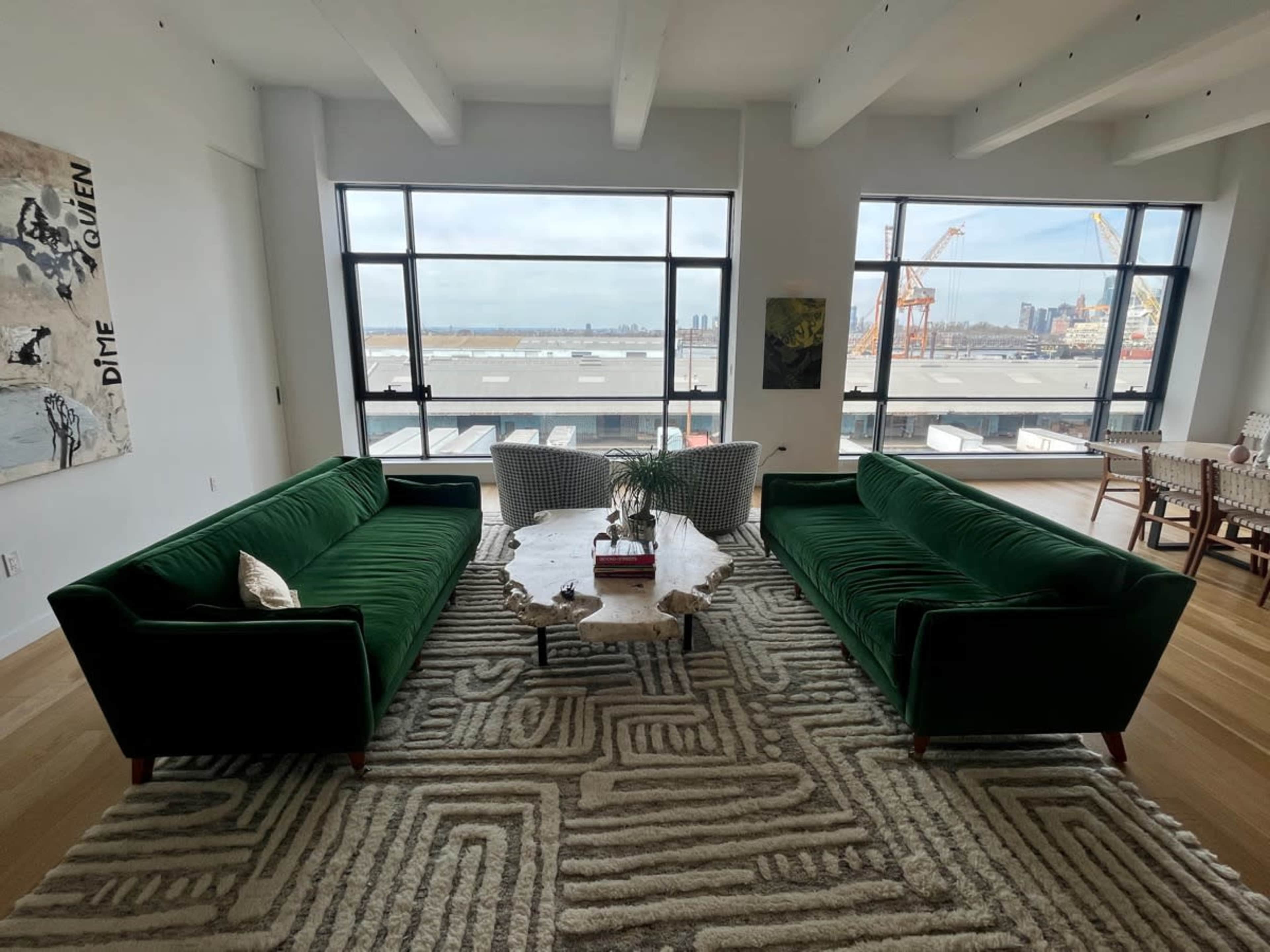 The scene depicts a spacious modern living area with two green velvet sofas facing a coffee table, surrounded by large windows displaying a view of a waterfront and distant city skyline.
