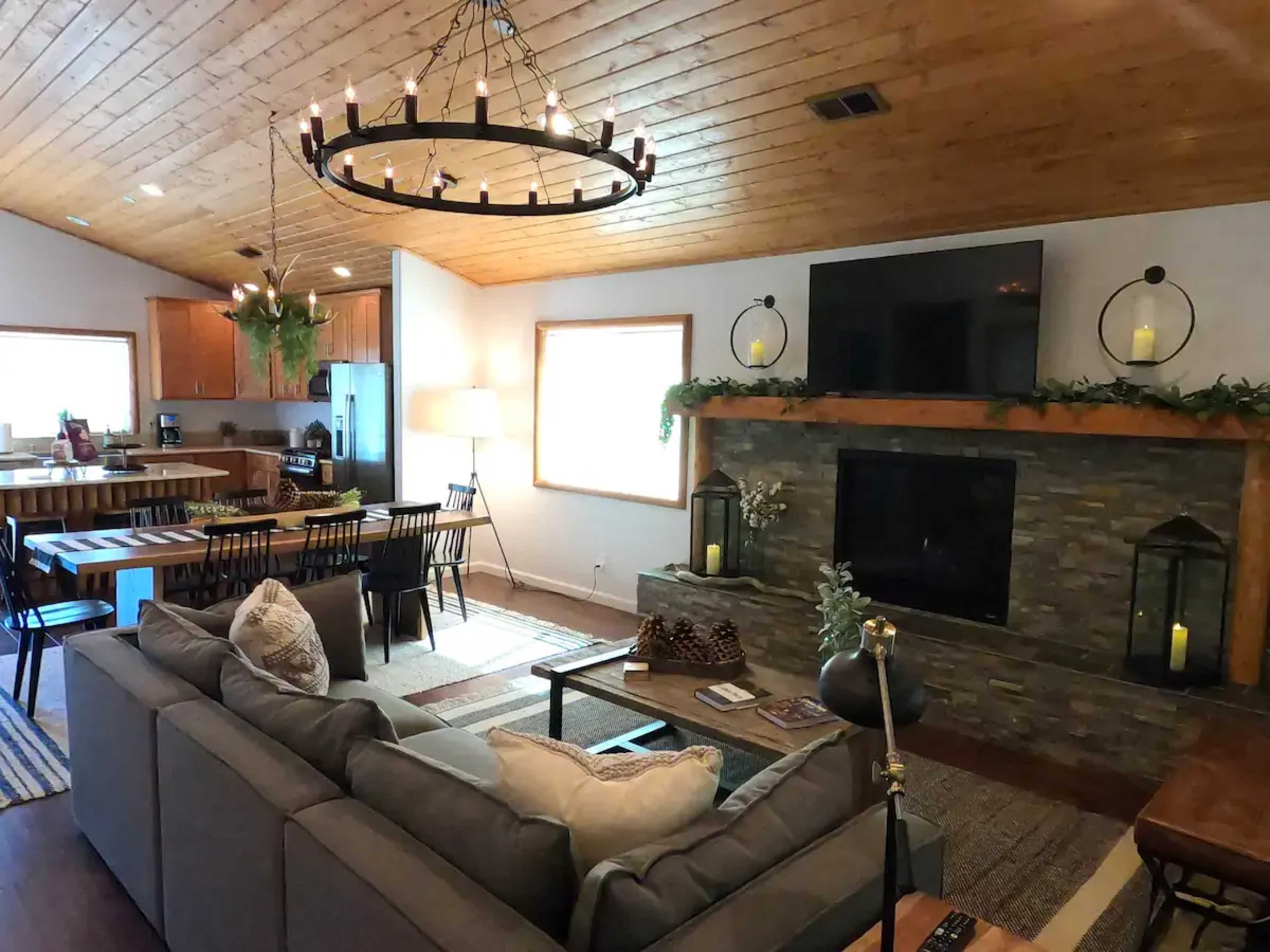The image shows a spacious living room with a gray sofa, a stone fireplace, a large flat-screen TV, and a dining area in the background.
