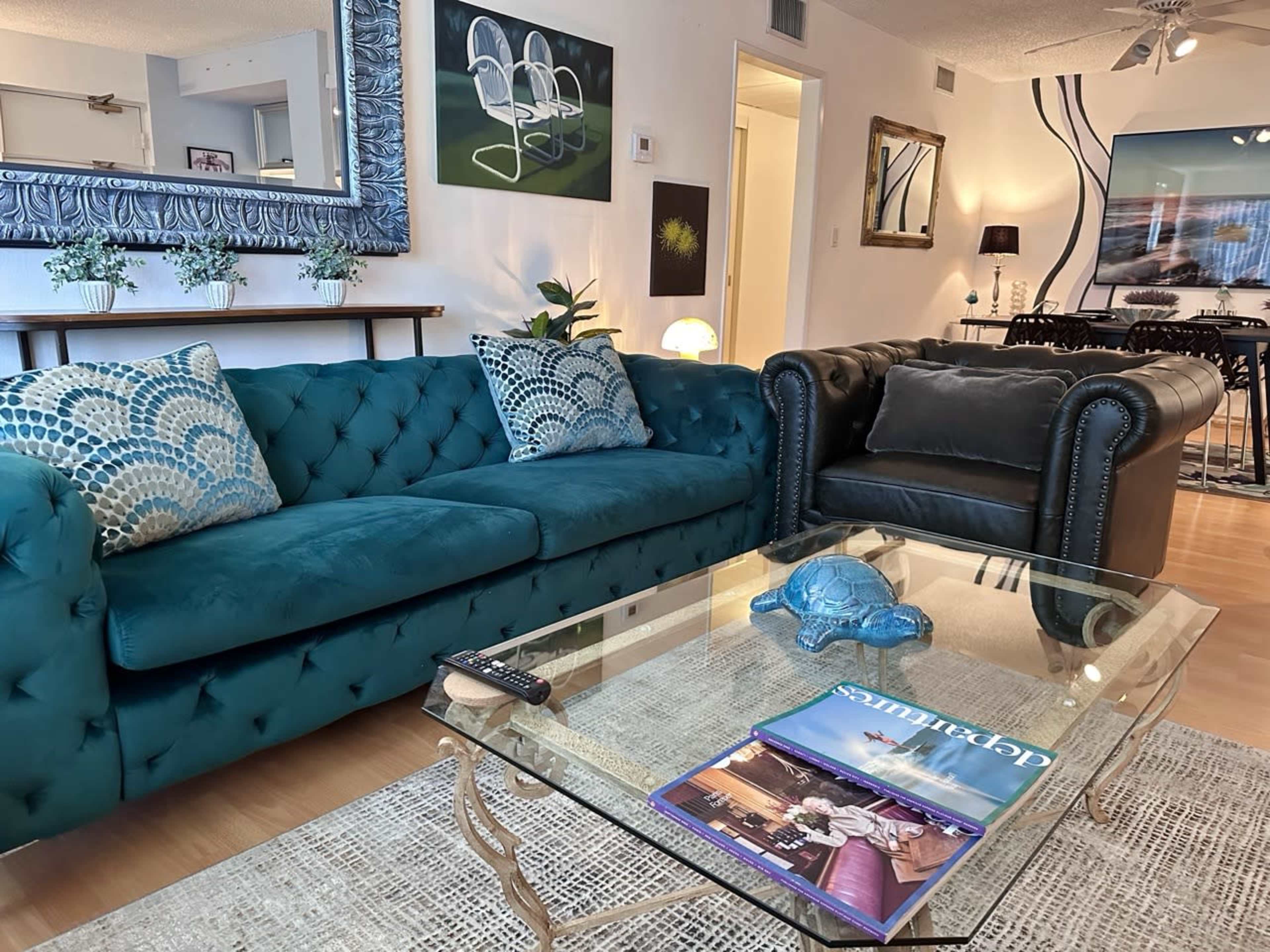 A teal tufted sofa and a black armchair are positioned around a glass coffee table in a styled living room featuring decorative artwork and modern furnishings.