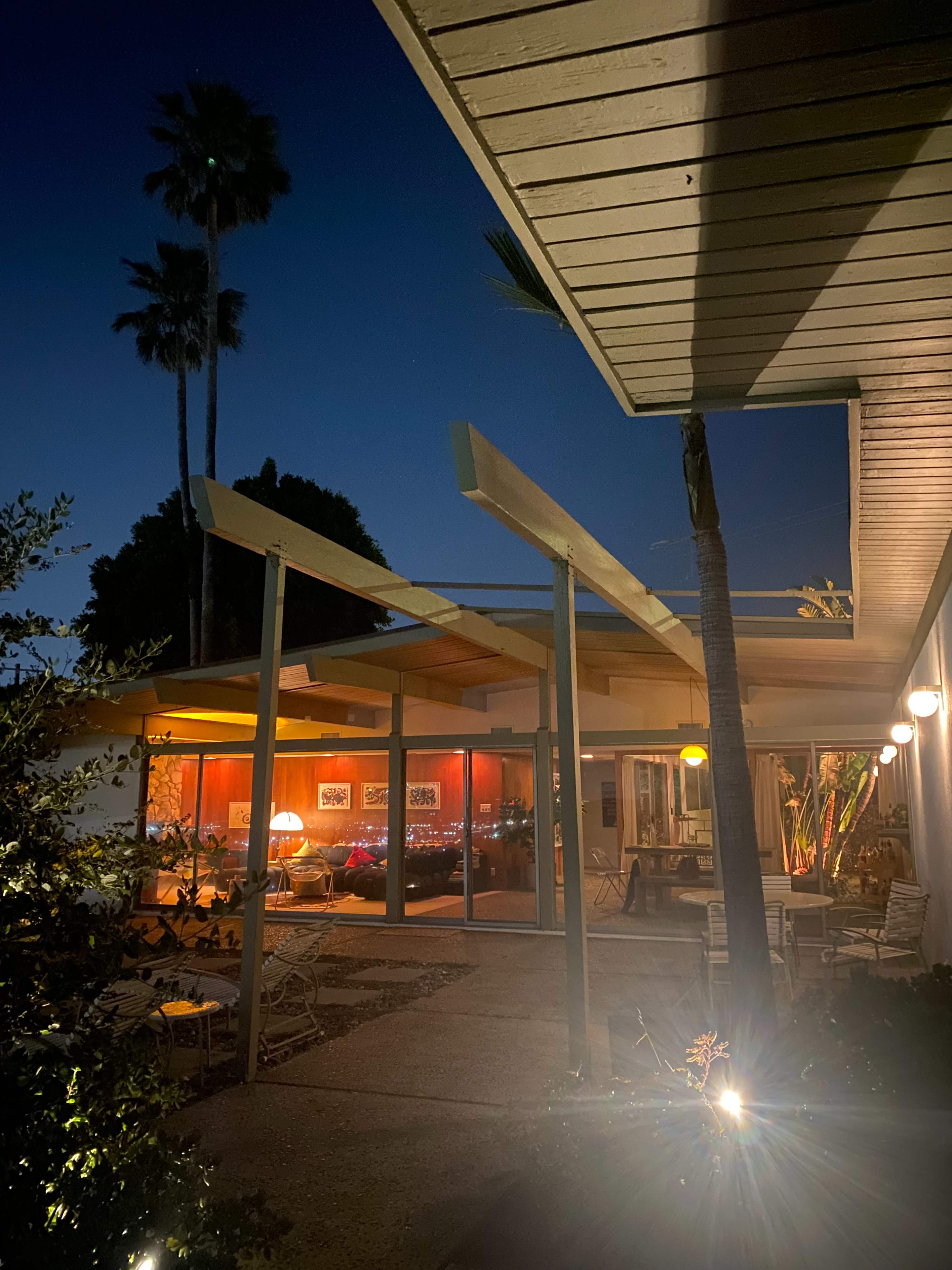 A modern architectural structure is illuminated at night, showcasing large windows that reveal an interior space with warm lighting and palm trees surrounding the entrance.