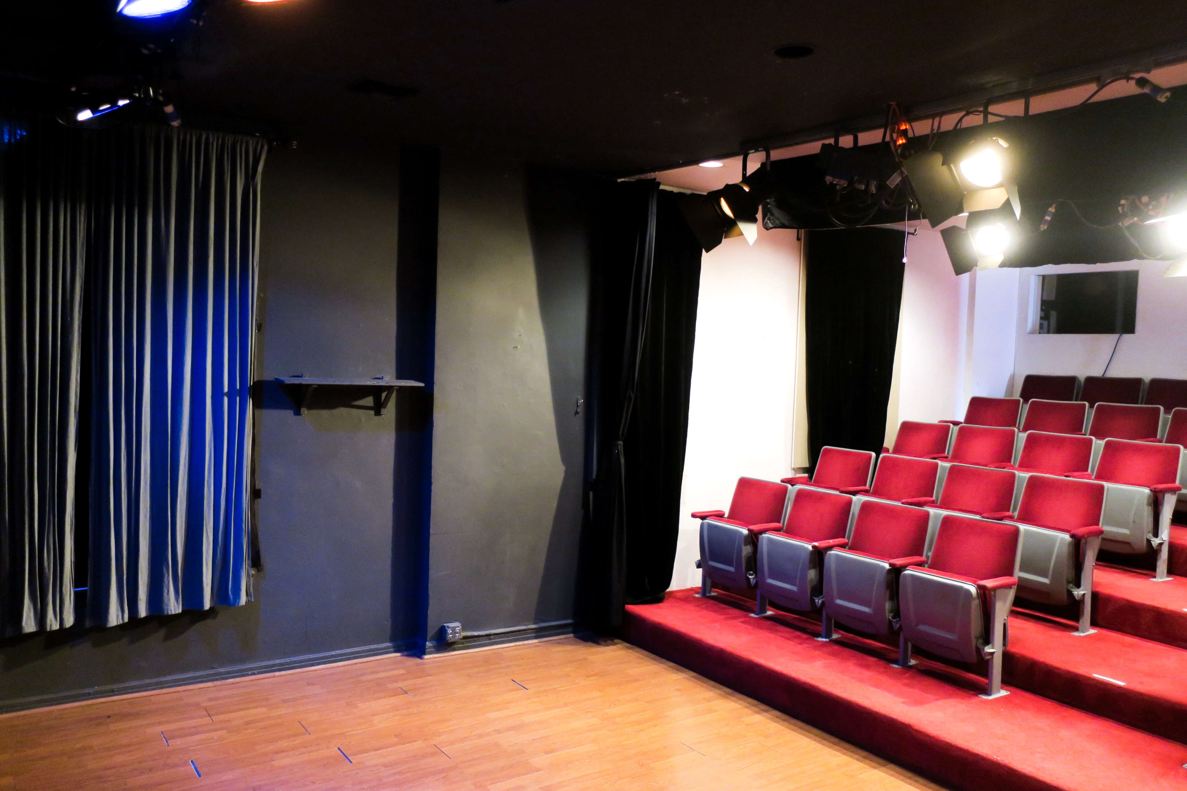 The image shows a small, empty theater with red seats facing a stage, flanked by curtains and lighting fixtures above.