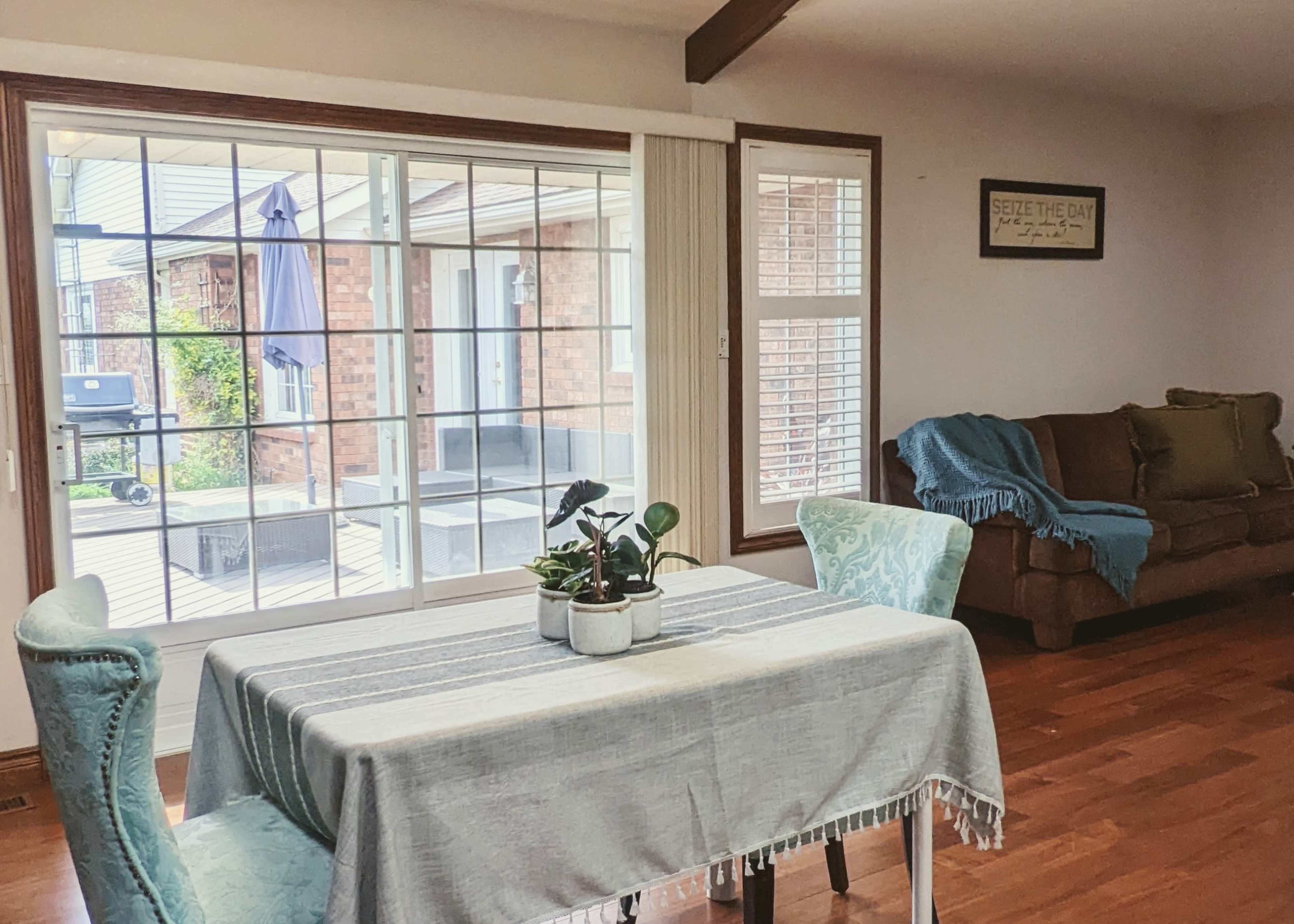 A table with decorative plants is positioned in front of a large window overlooking a patio area, with a couch nearby.