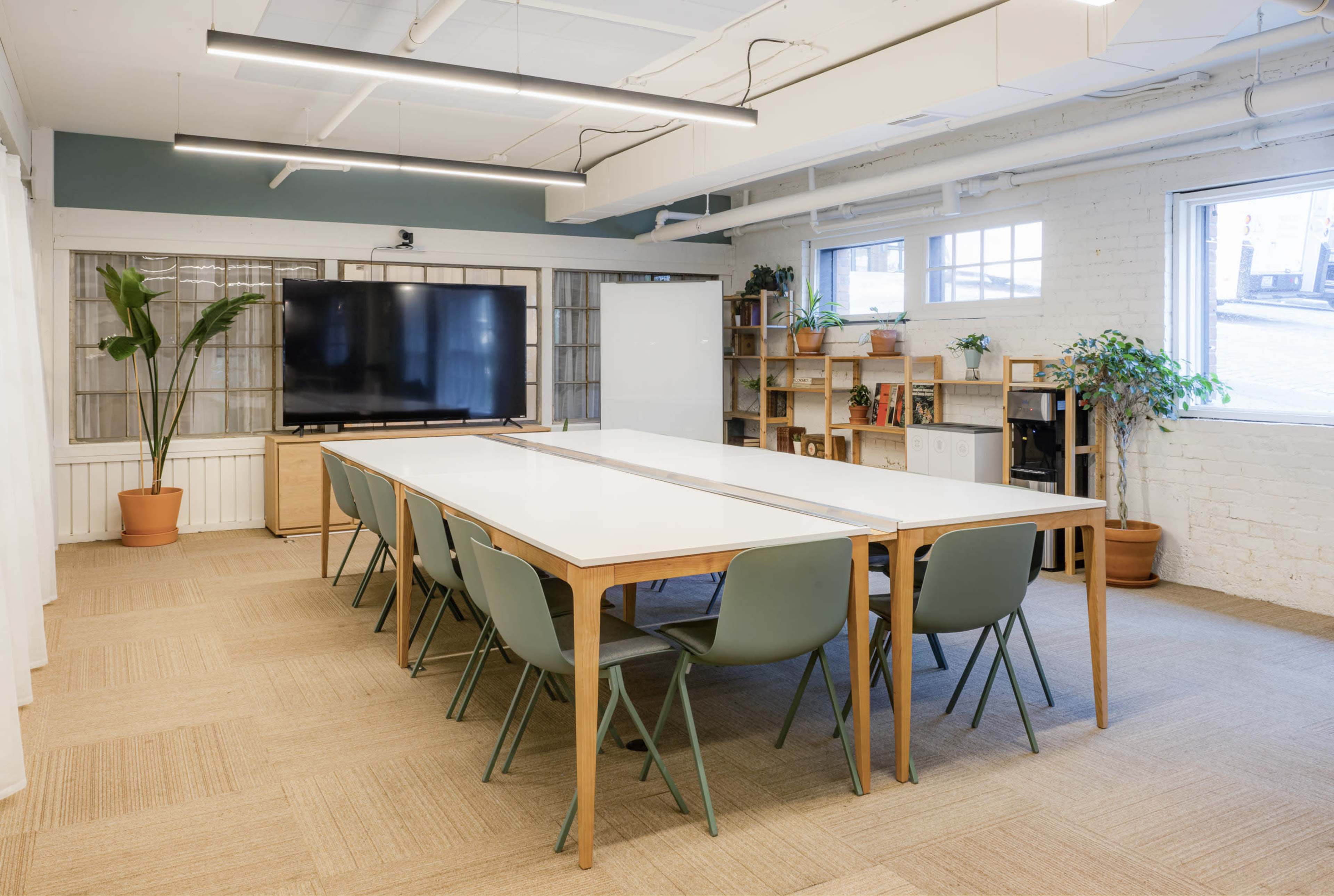 A bright meeting room features a long white table surrounded by green chairs, with a television on the wall and shelves displaying plants.