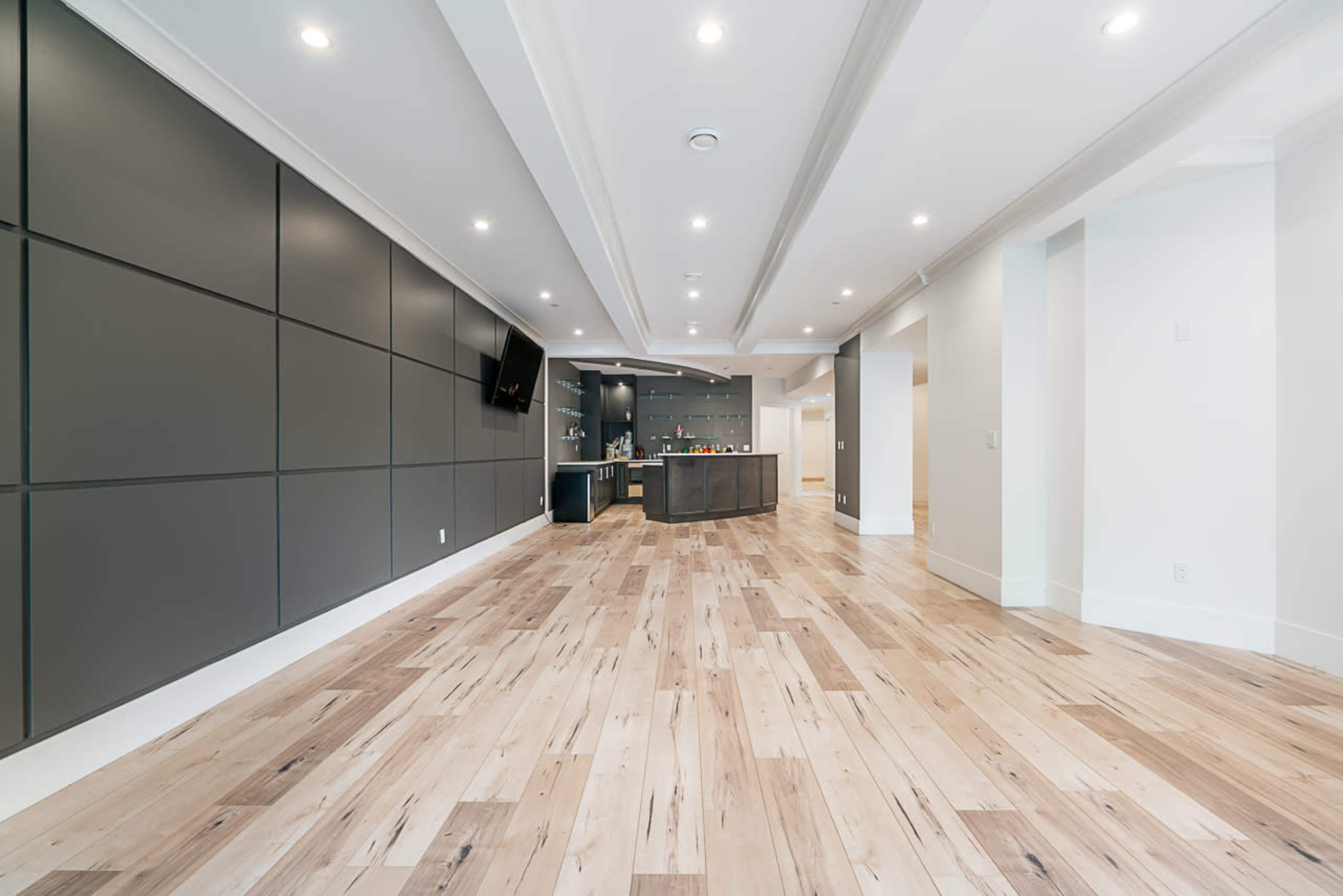 A spacious interior room with wooden flooring, gray panel walls, and a kitchen area featuring a dark countertop and shelves.