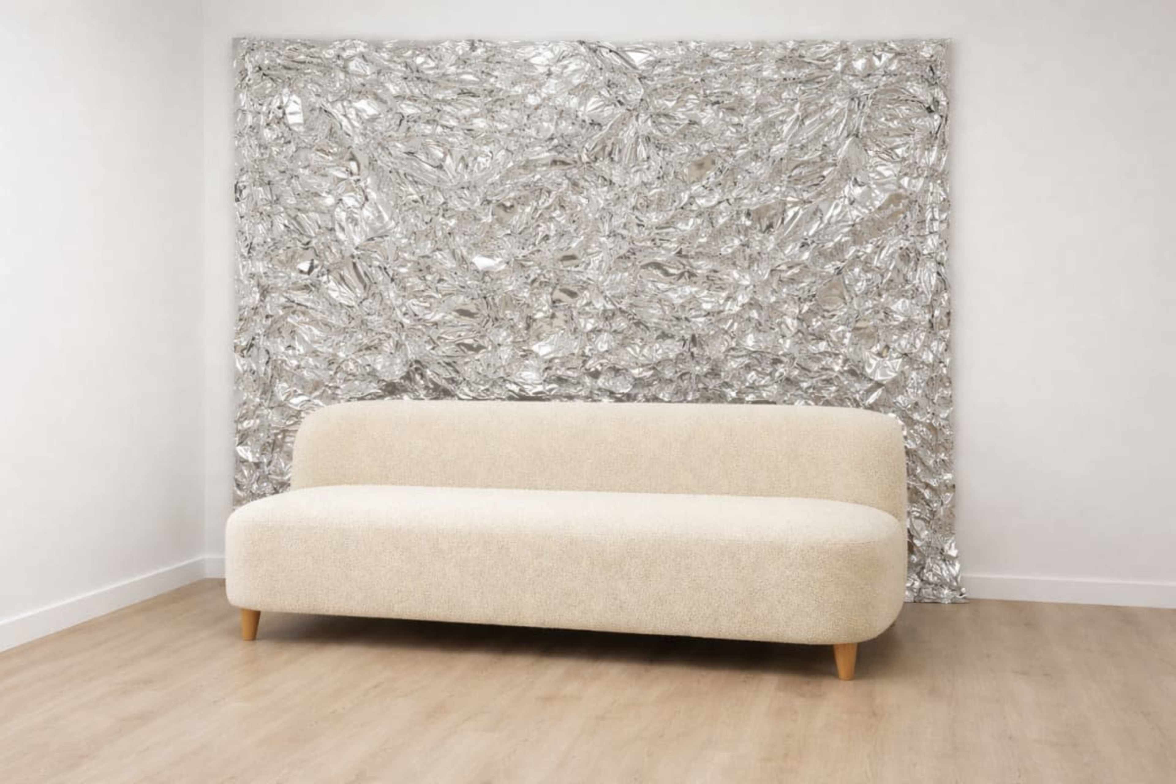 A beige, curved sofa sits in front of a textured silver foil wall in a simple room with wooden flooring.