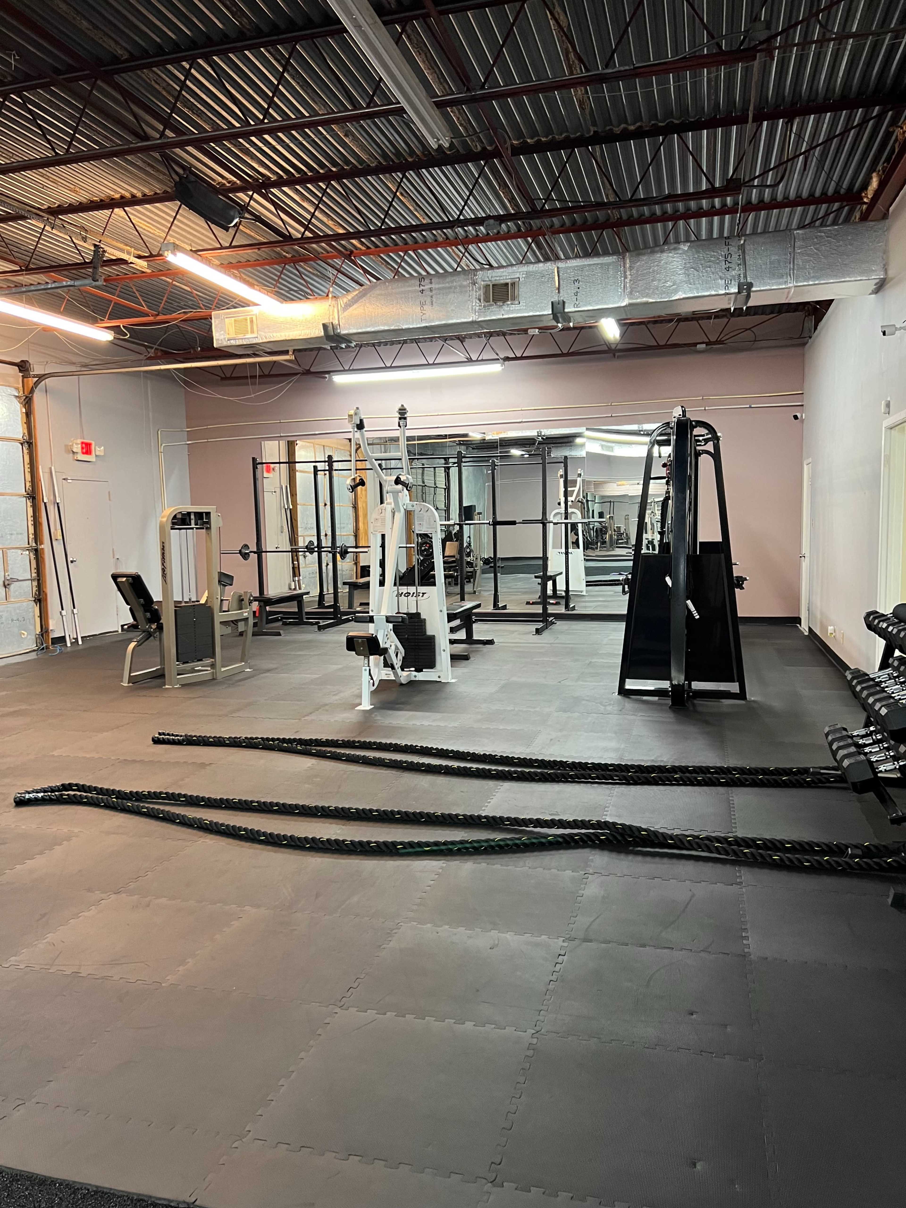A well-equipped gym with various weight machines, dumbbells, and training ropes arranged on a black rubber floor.