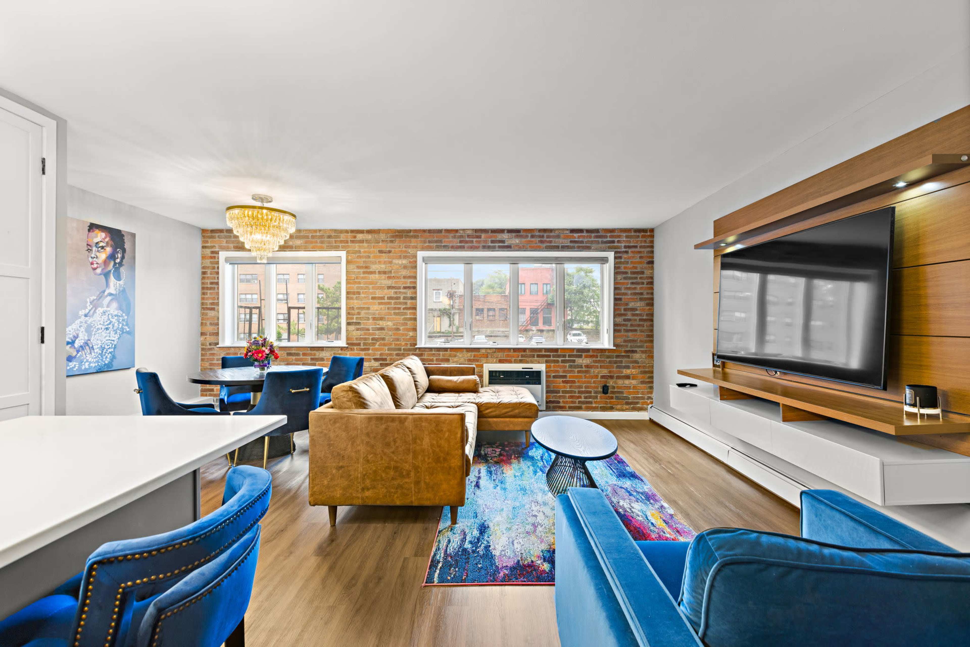 A modern living room features a seating area with brown and blue couches, a large TV mounted on a wooden wall unit, and a dining space with blue chairs and a table.