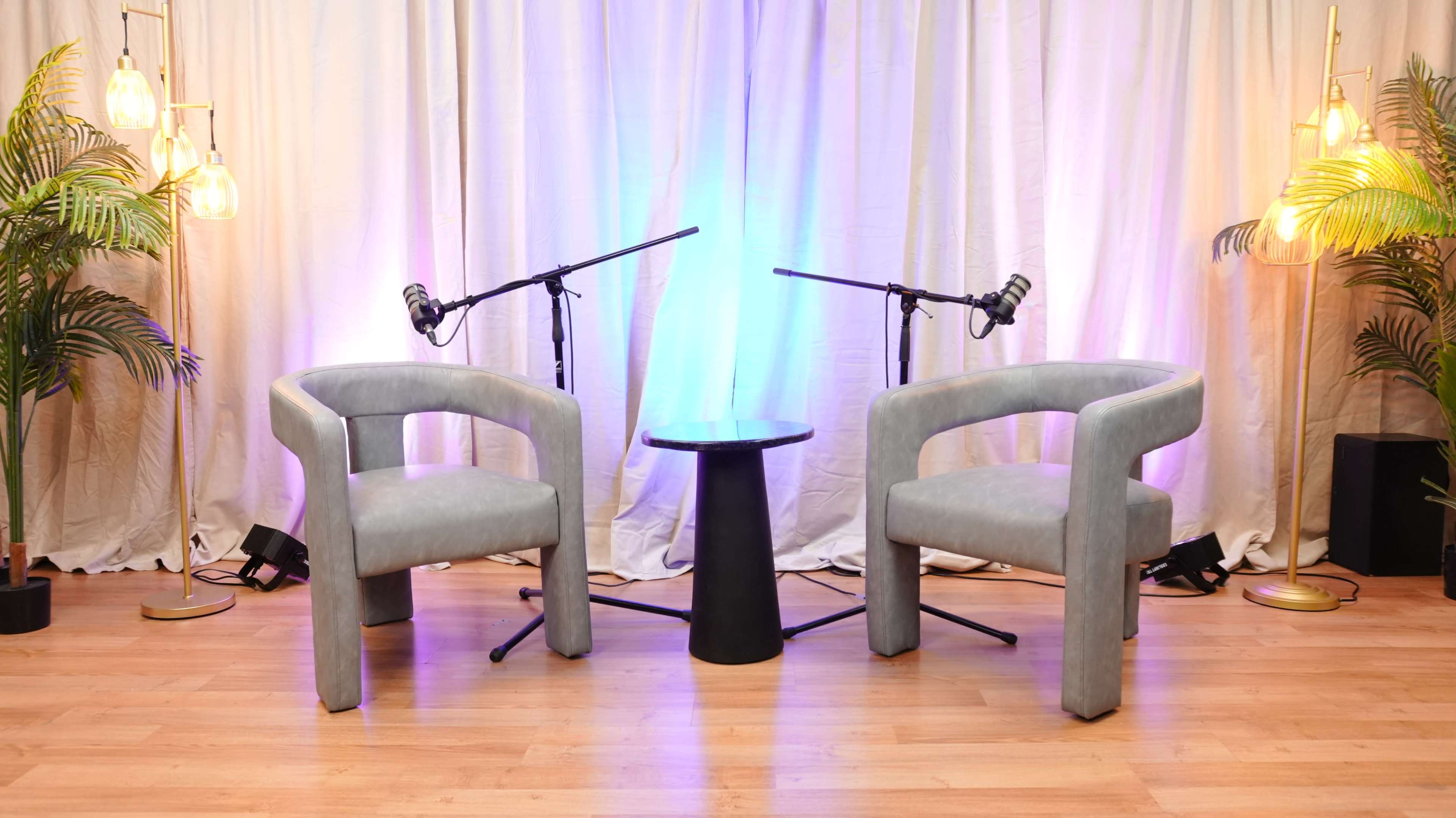 The image shows a studio setup featuring two light gray chairs, a black table in between, and two microphones on stands, with a softly lit background in pastel colors.
