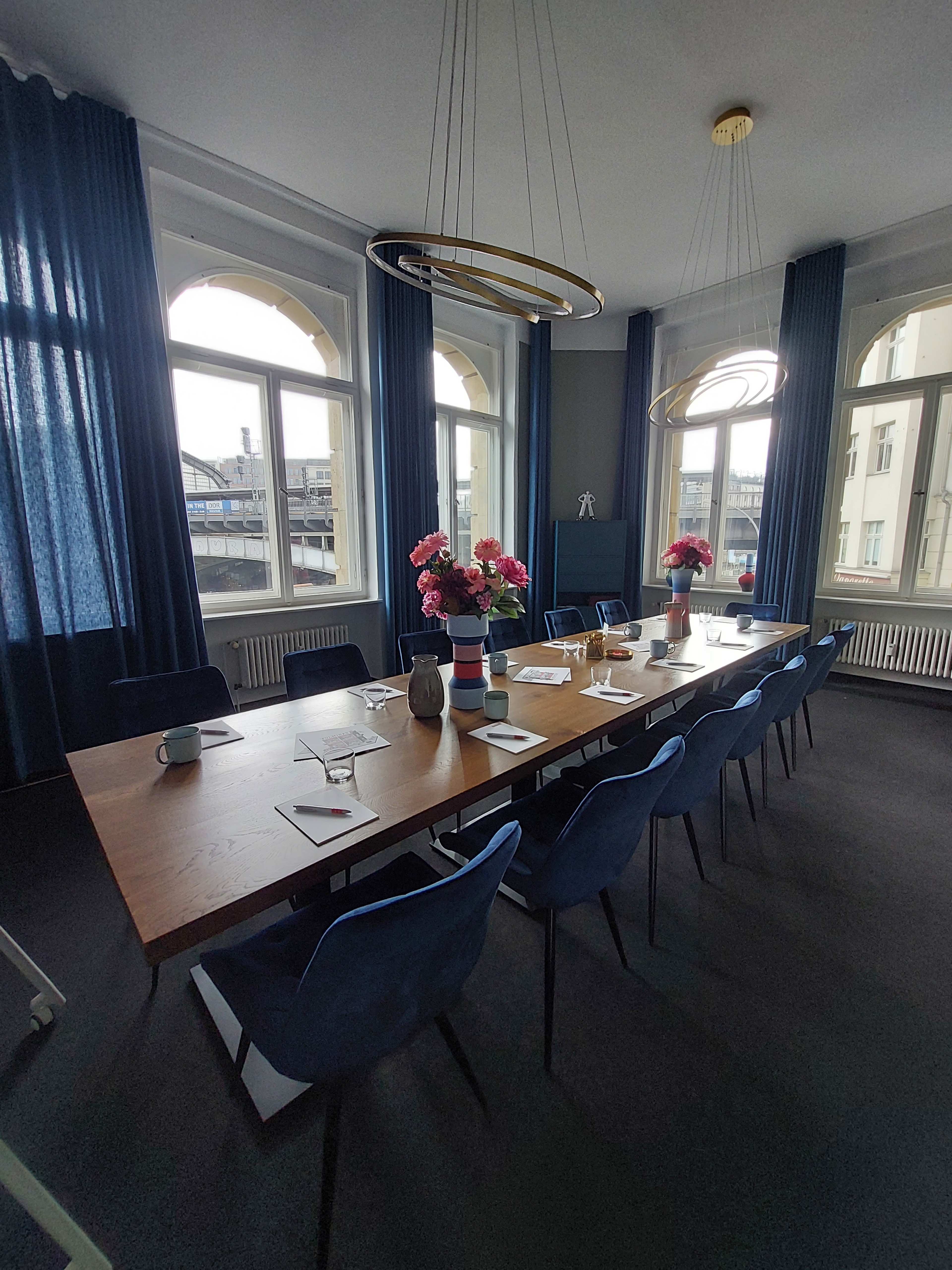 A modern conference room features a large wooden table surrounded by blue chairs, with large windows and decorative flower arrangements.