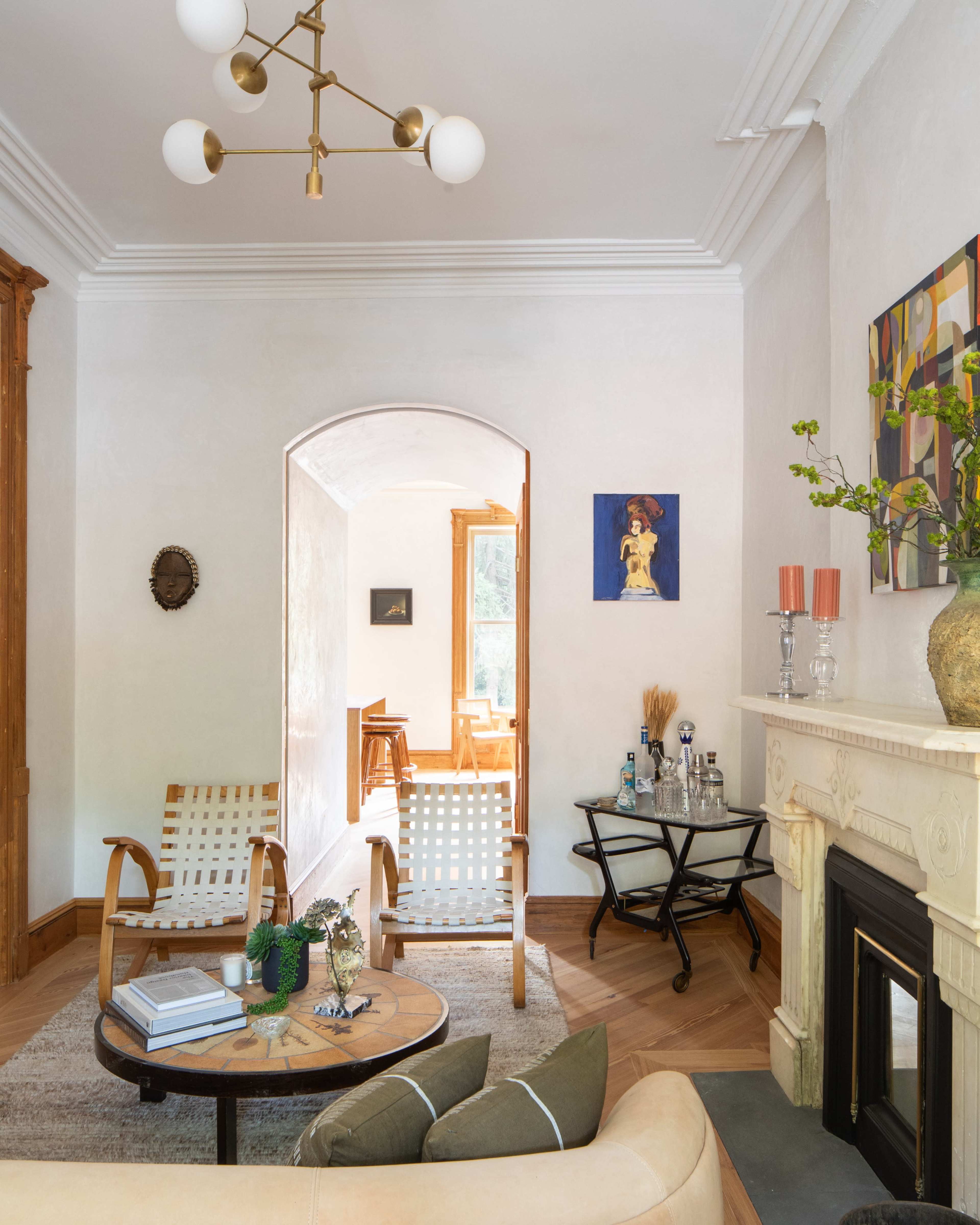 The image depicts a cozy living room featuring a beige sofa, wooden chairs, a round coffee table, and a decorative fireplace, along with an archway leading to another room.
