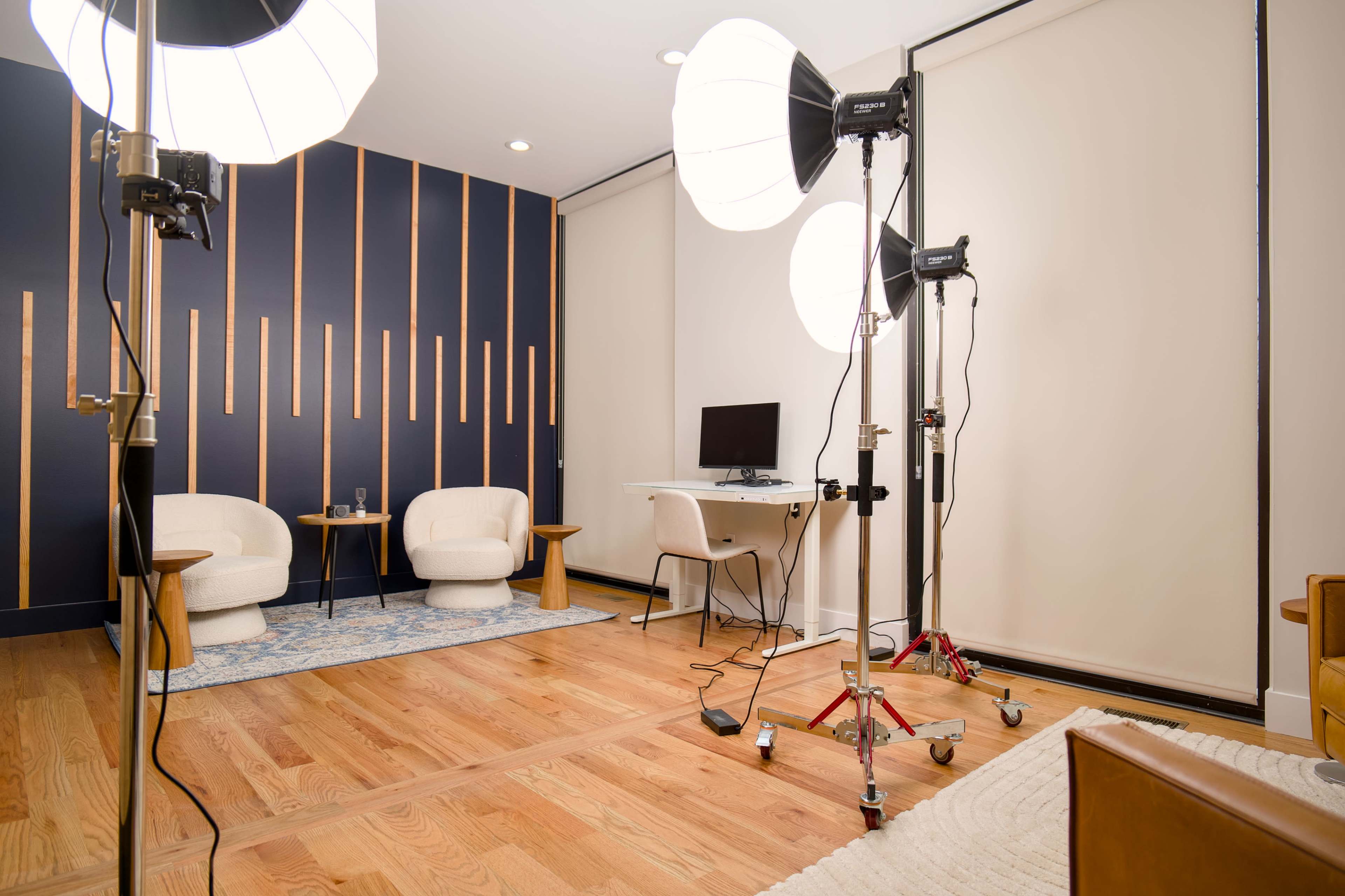 The image depicts a modern studio setup with two lighting stands, a small desk with a computer, and two white armchairs against a dark blue wall.