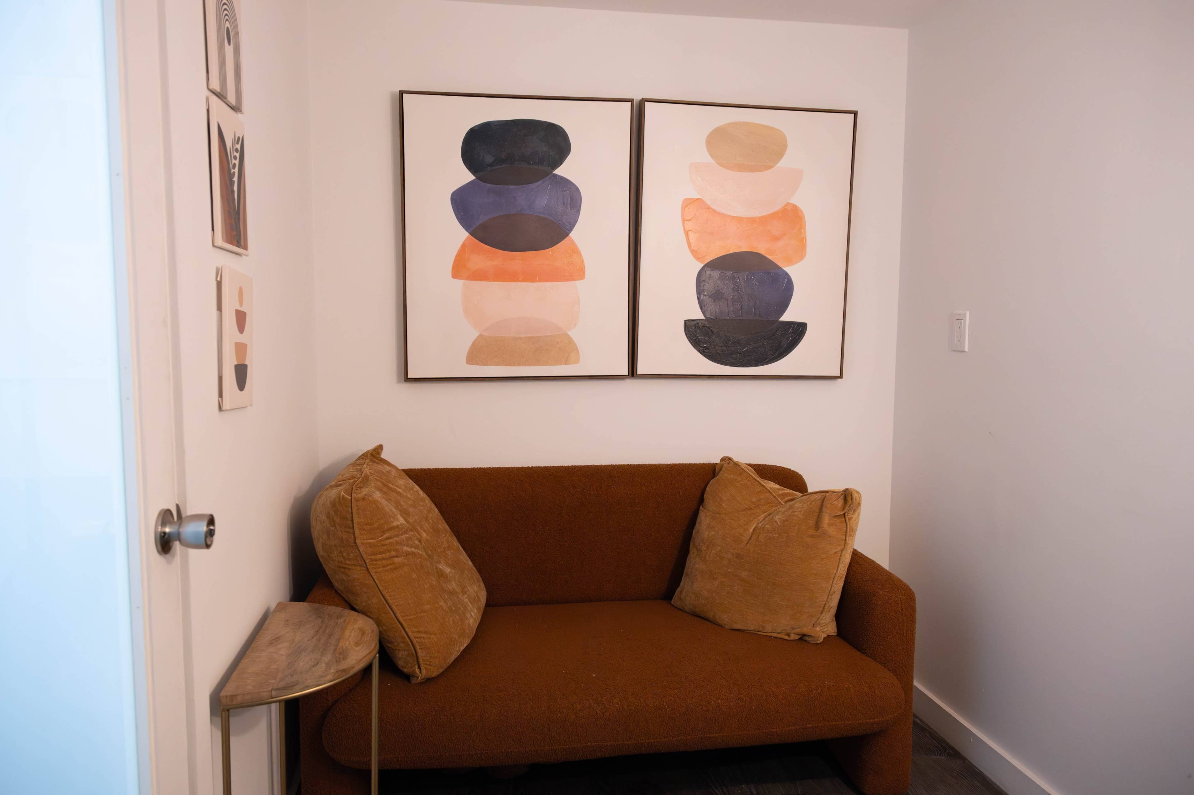The image shows a small corner of a room featuring a brown sofa with two decorative pillows and two framed abstract art pieces on the wall above it.