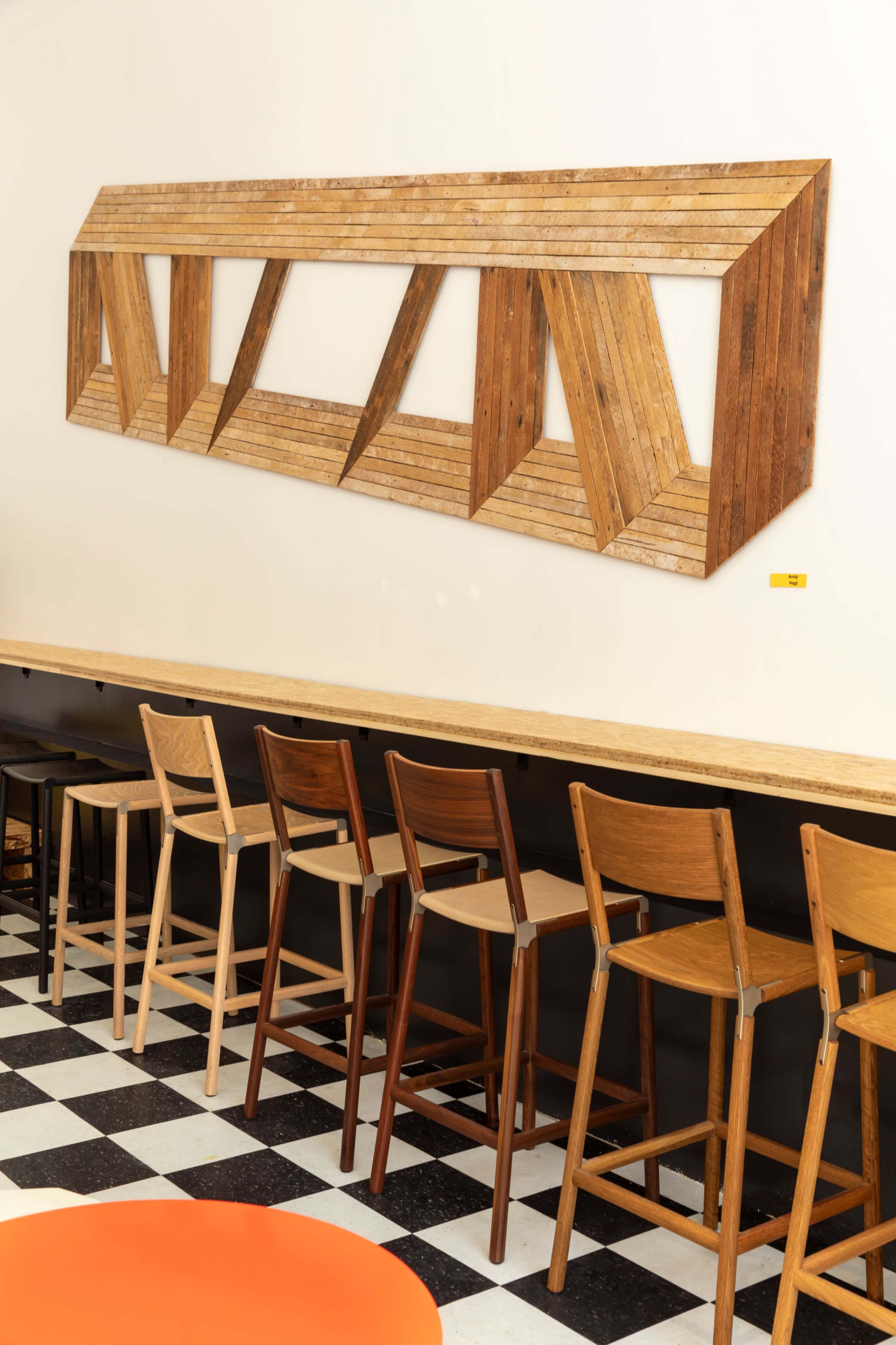 The image shows a wooden wall installation with a geometric design above a row of various wooden bar stools in a café setting.
