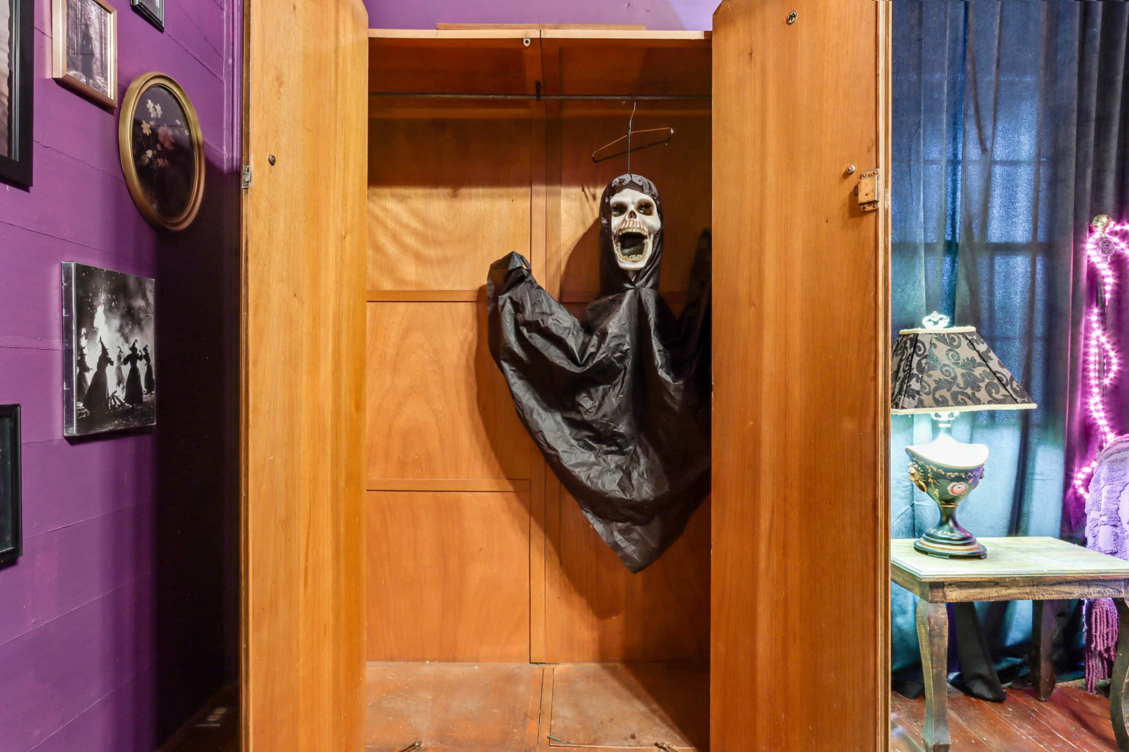 A wooden wardrobe with a hanging black cloak and a skull mask is positioned against a purple wall, alongside framed photographs and a lamp.