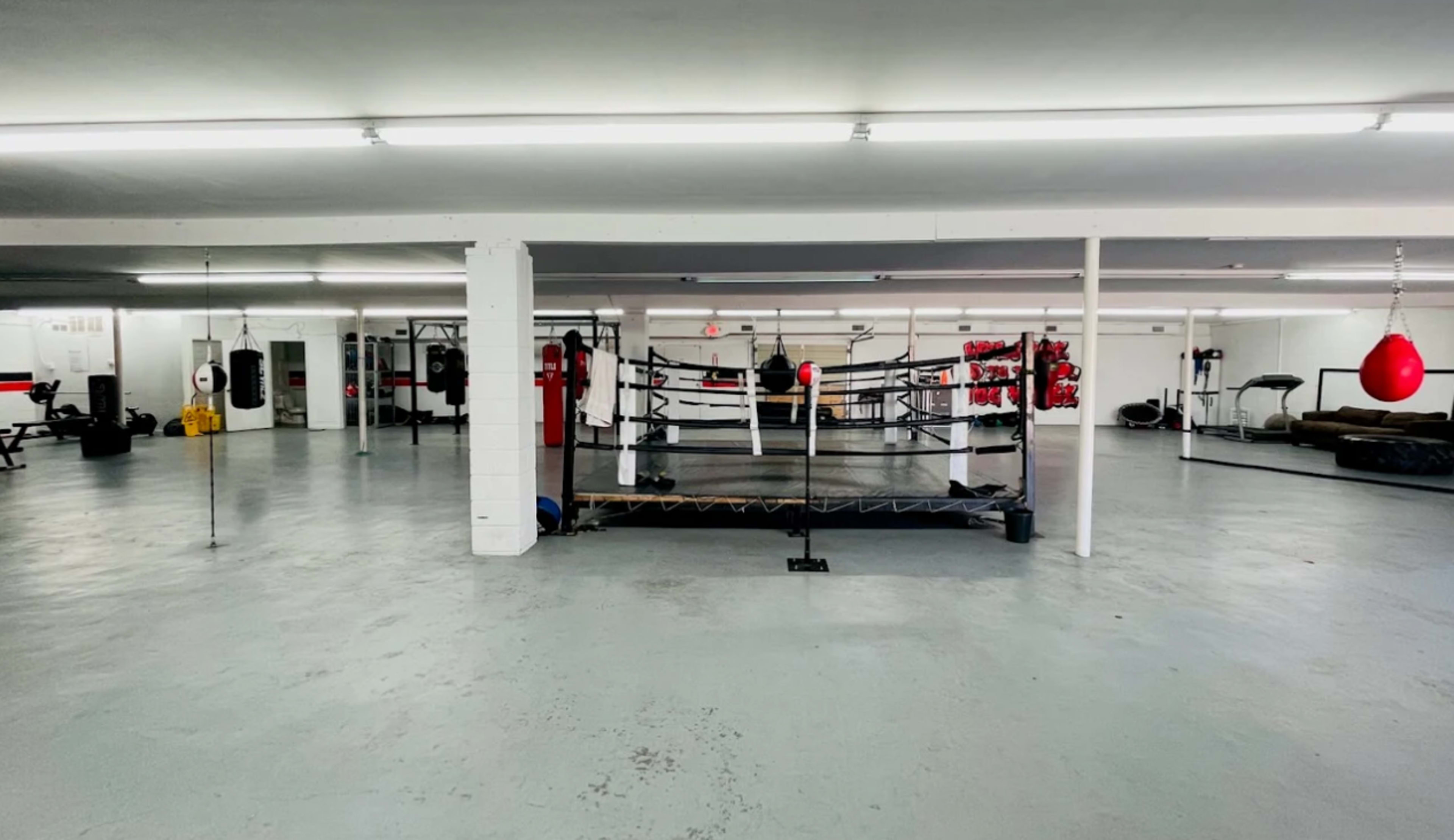 The image shows a spacious, well-lit boxing gym with a boxing ring in the center surrounded by various training equipment.