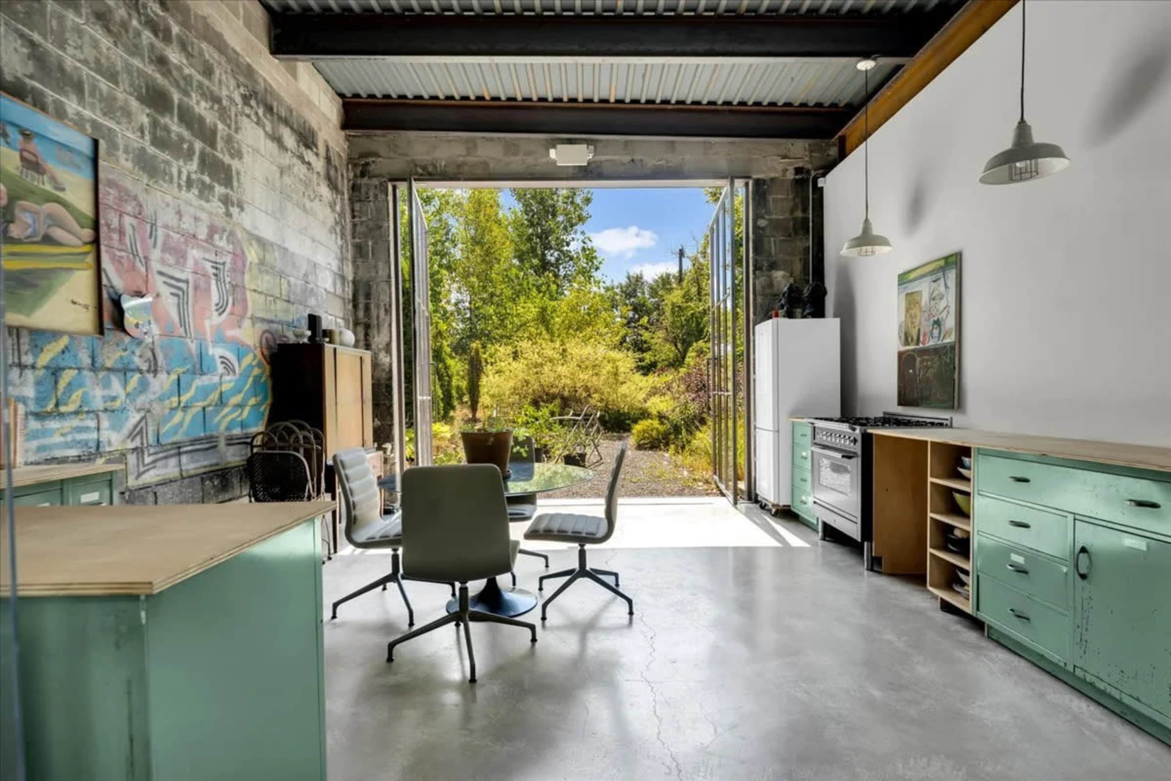 The image shows an industrial-style interior with a kitchen area, a dining table with chairs, and a large open door leading to a landscaped outdoor area.