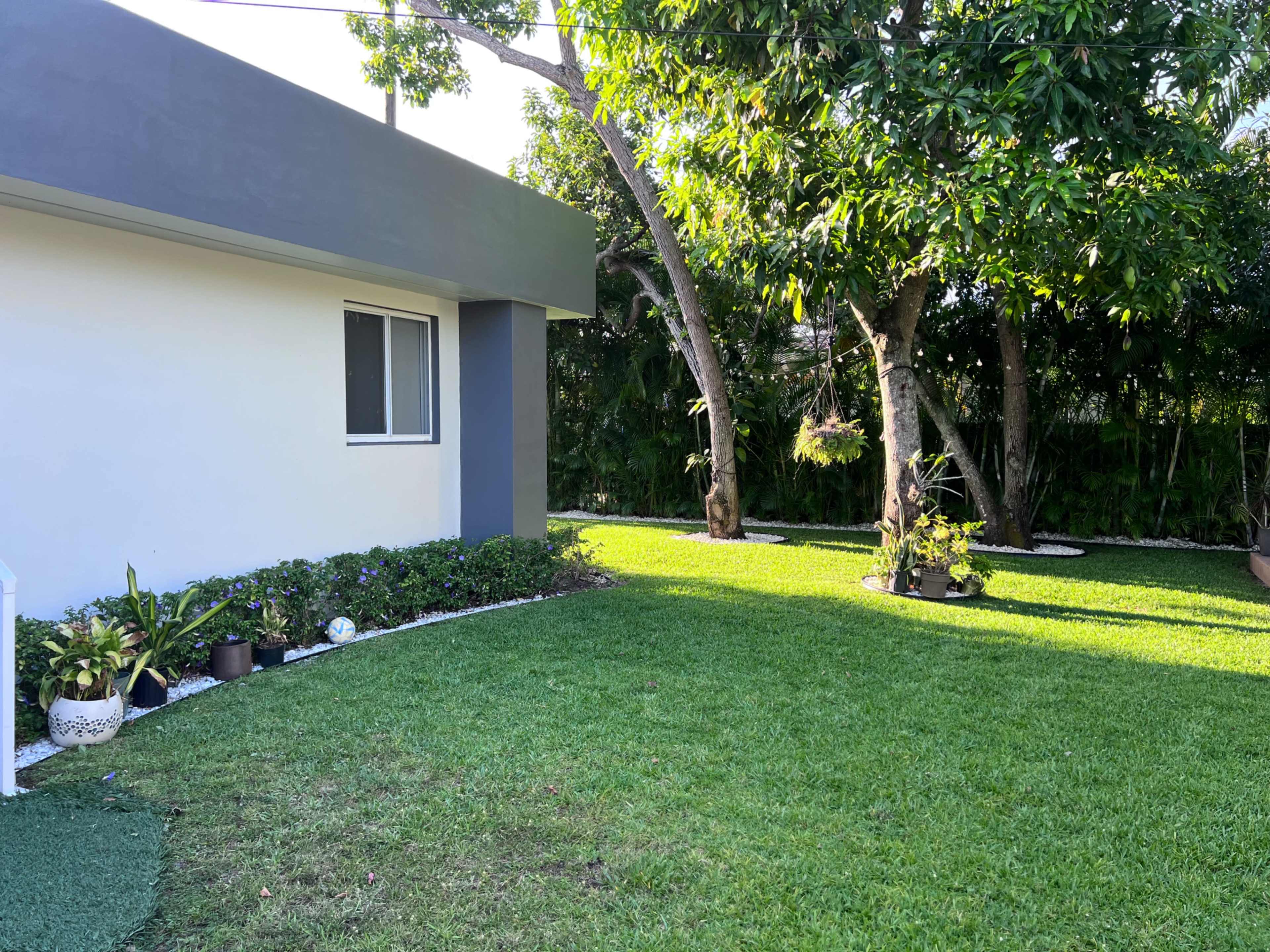 The image shows a well-maintained grassy yard with a white and gray house on one side and trees with potted plants on the other.