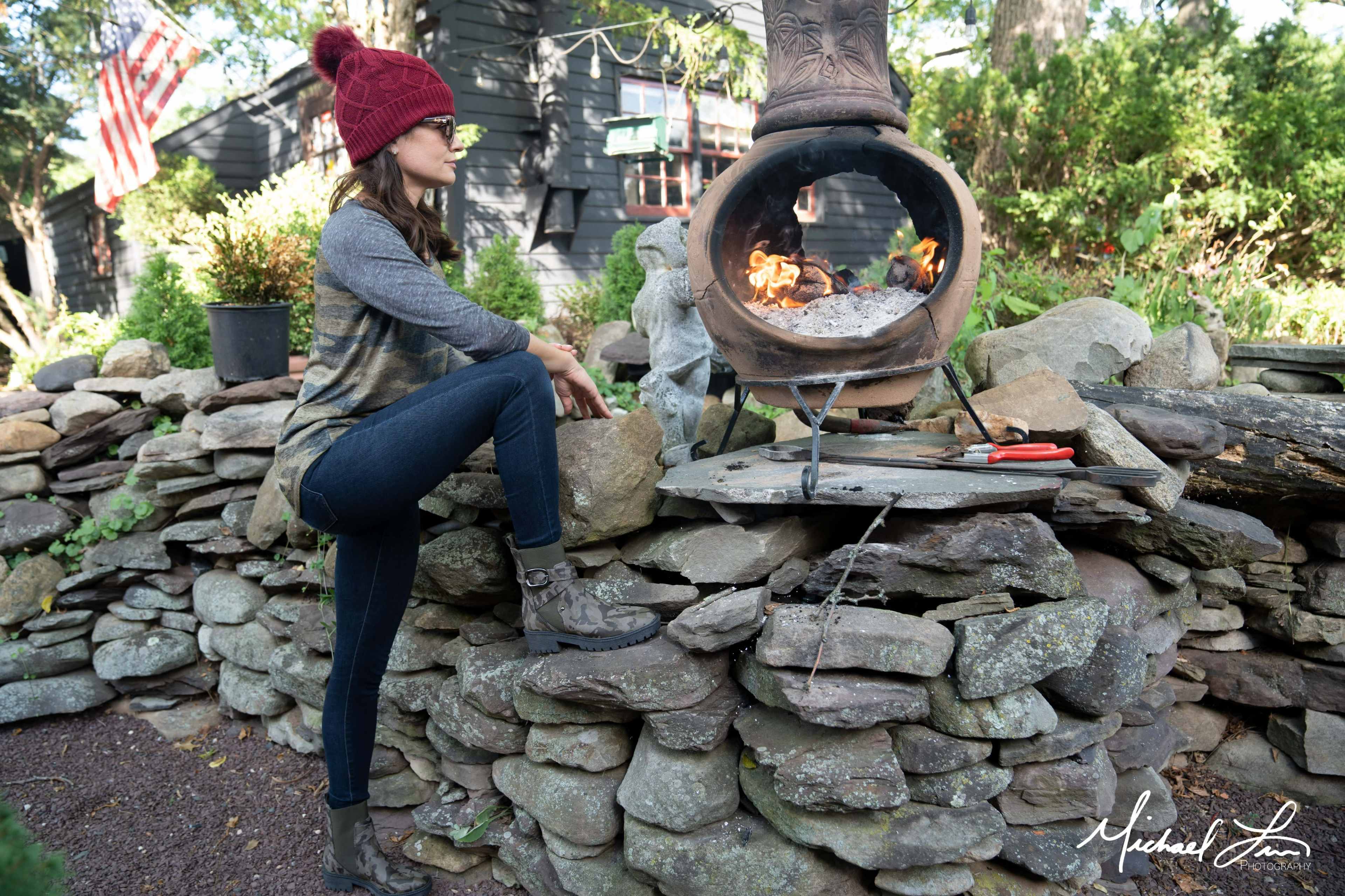 A person in a red beanie sits on a stone wall next to a fire burning in a clay chiminea.